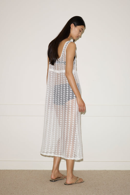 A woman with long dark hair stands sideways against a plain wall, wearing the 0 Thread Dress in Off White over a black bikini and beige sandals.
