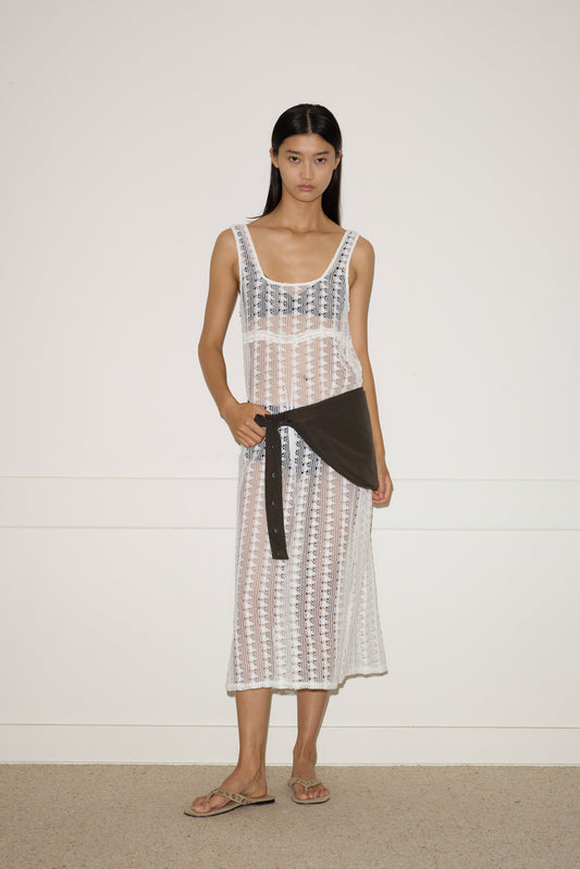 A woman with straight, long dark hair and a neutral expression stands against a plain wall, wearing the 0 Thread Dress - Off White over a black bikini, paired with a dark shirt at her waist and beige sandals.