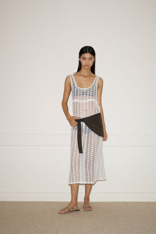 A woman stands against a plain wall wearing a sleeveless white crocheted dress over a black bikini, sandals, a black long-sleeve shirt tied at her waist, and carrying the Press Stud Saddle Bag in Dark Chocolate.