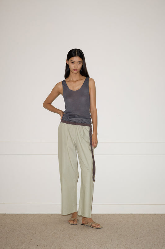 A woman with straight dark hair stands indoors against a plain white wall, wearing a sleeveless dark gray top, loose light beige organic cotton pants secured with the 0 Plaited Belt in Chocolate, and sandals. One hand rests on her hip while the other hangs by her side.