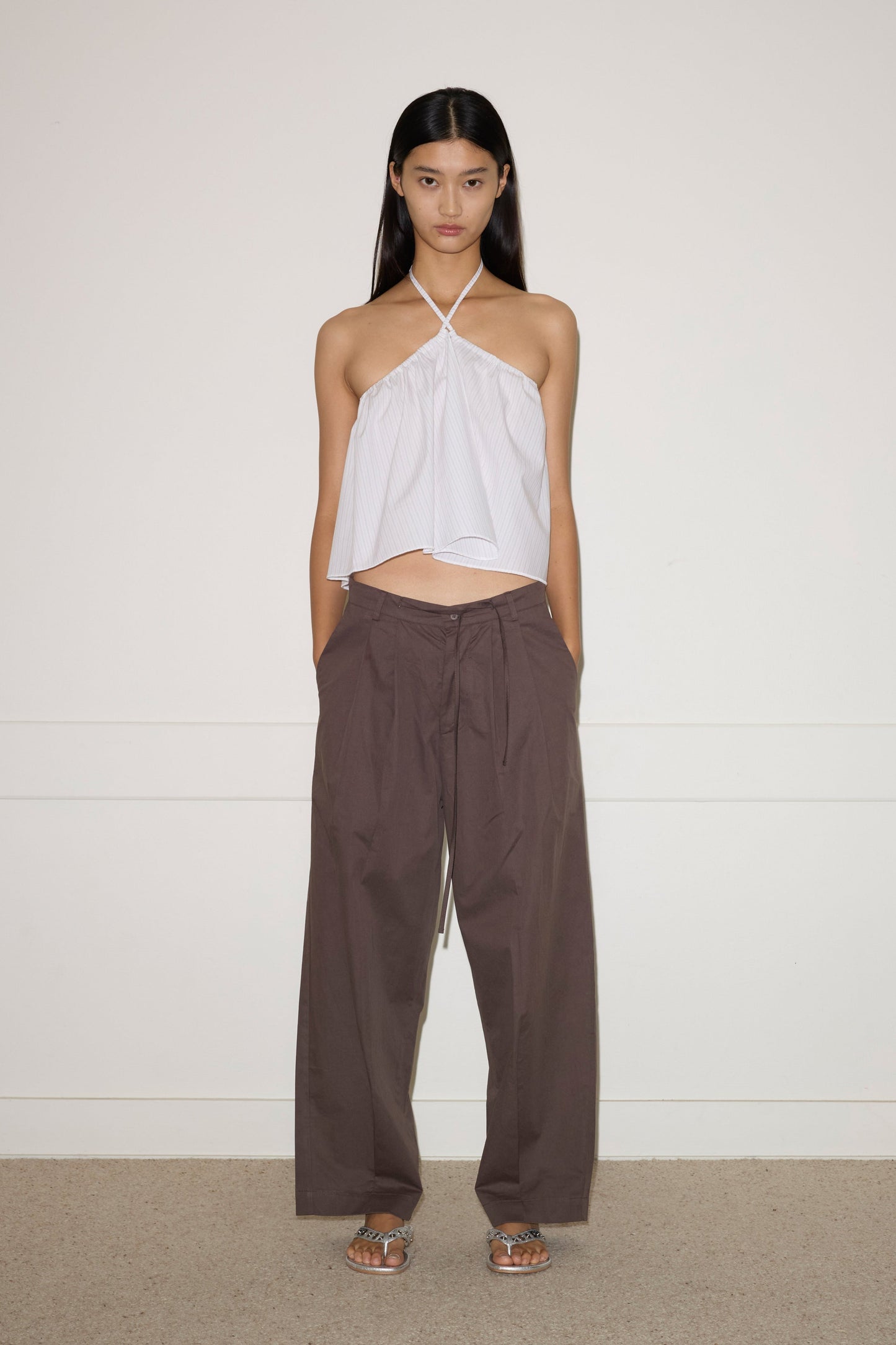 A woman stands against a plain wall, wearing the Turn Pant - Cocoa by 0, featuring relaxed tailoring, organic cotton fabric, and an adjustable drawcord waist. She pairs them with a white halter-neck top and silver sandals. Her hair is straight; her expression neutral.
