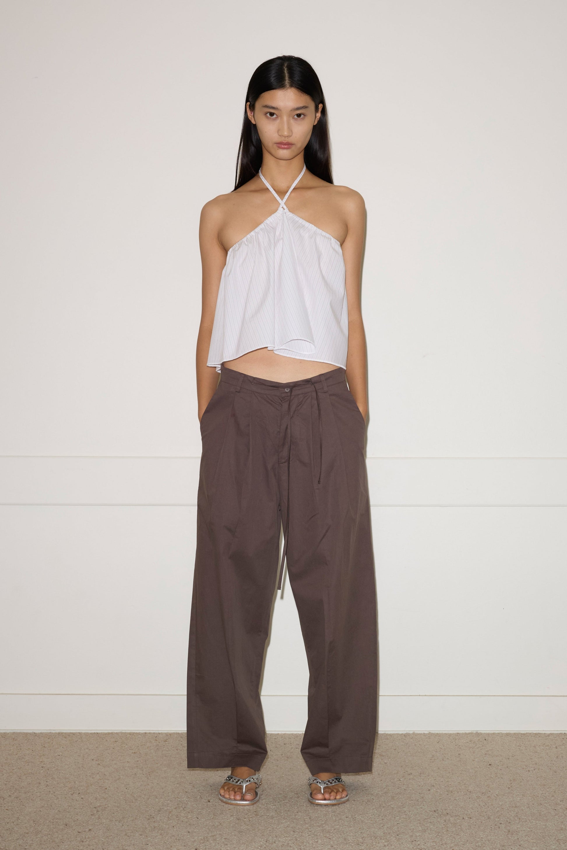 A woman stands against a plain wall, wearing the Turn Pant - Cocoa by 0, featuring relaxed tailoring, organic cotton fabric, and an adjustable drawcord waist. She pairs them with a white halter-neck top and silver sandals. Her hair is straight; her expression neutral.