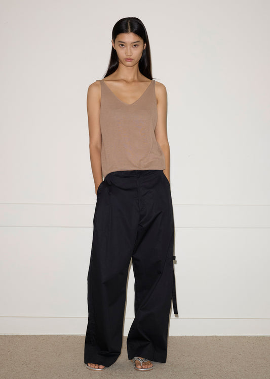A person with long dark hair stands against a white wall, wearing the Tab Pant in Black, a loose organic cotton sleeveless top, and open-toe shoes. Their hands are in their pockets and they have a neutral expression.
