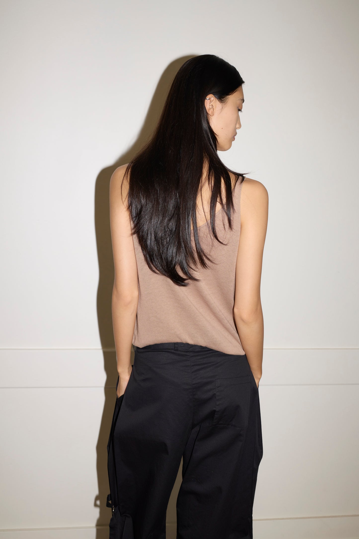 A woman with long, straight dark hair faces away from the camera, wearing a beige sleeveless top and the Tab Pant - Black against a plain light-colored wall.