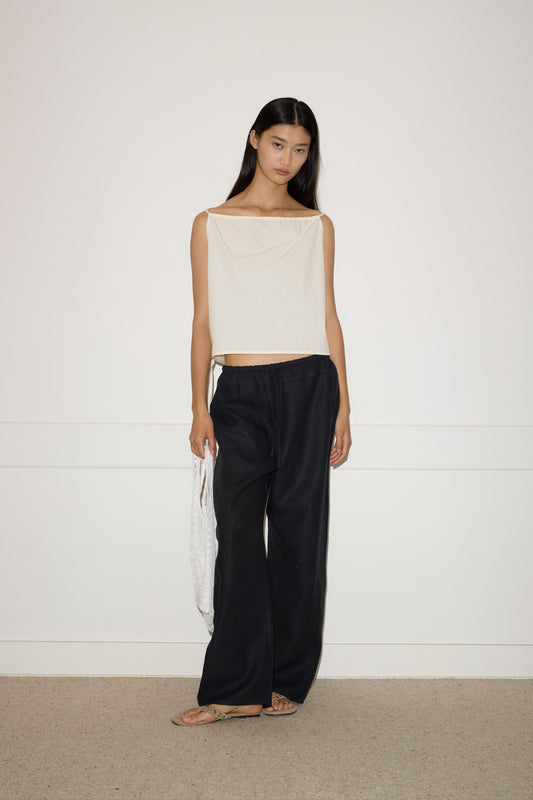 A woman with long dark hair stands indoors by a white wall, dressed in the Simple Top - Butter White by Deiji Studios, loose black pants, and sandals, while holding a white net bag.