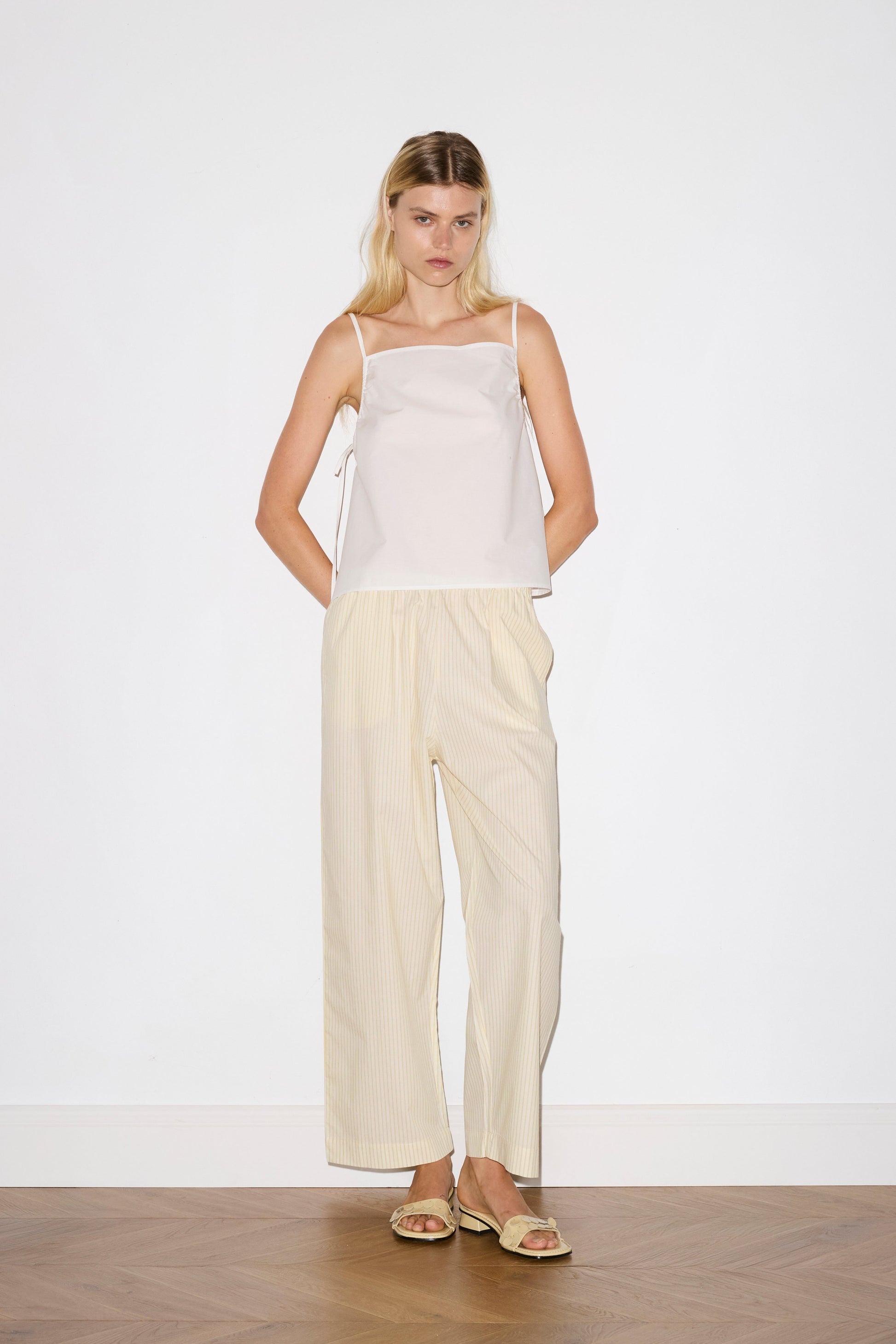 blonde female model wears a white lightweight top featuring fixed gathered armholes and right hand side armhole keyhole and ties, paired with a butter yellow striped ankle length pant features a soft minimal elastic waist with side seam pockets in a straight leg