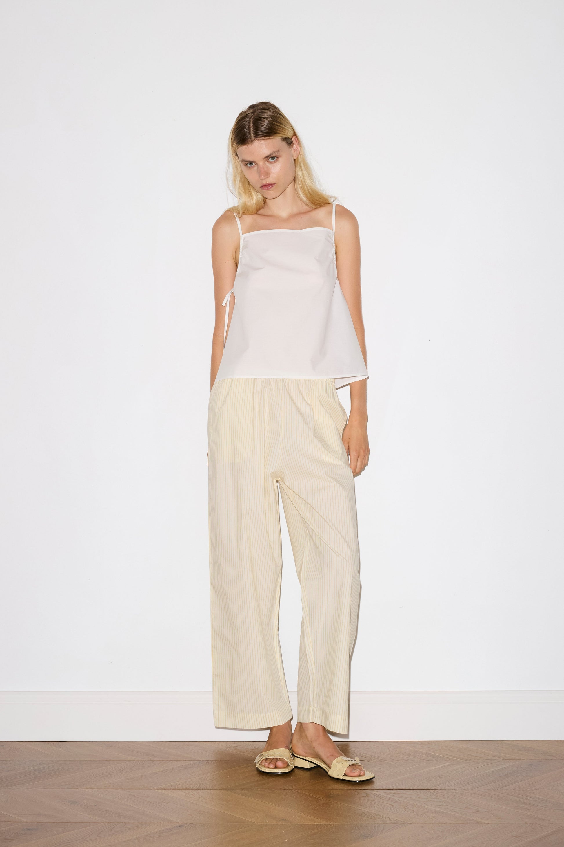 blonde female model wears a white lightweight top featuring fixed gathered armholes and right hand side armhole keyhole and ties, paired with a butter yellow striped ankle length pant features a soft minimal elastic waist with side seam pockets in a straight leg