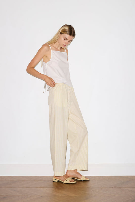 blonde female model wears a white lightweight top featuring fixed gathered armholes and right hand side armhole keyhole and ties, paired with a butter yellow striped ankle length pant features a soft minimal elastic waist with side seam pockets in a straight leg