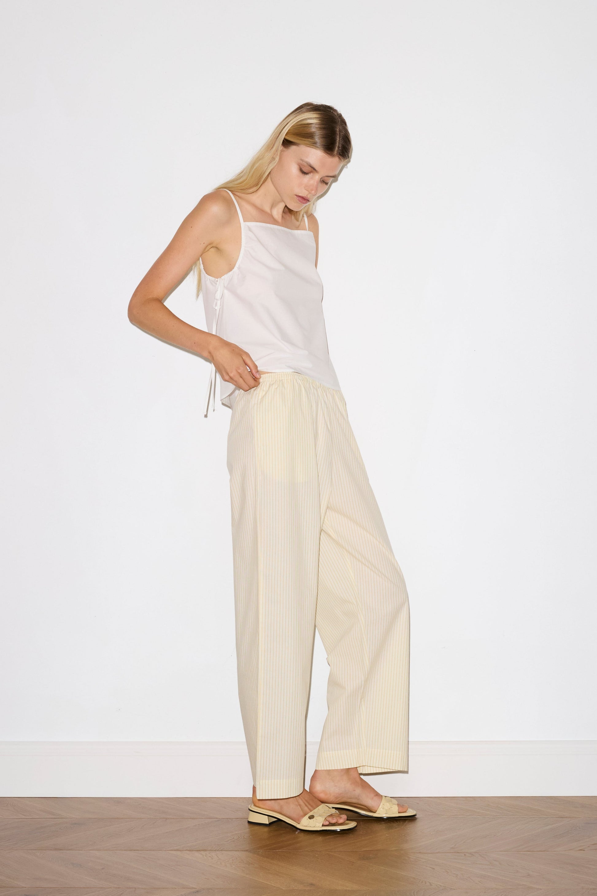 blonde female model wears a white lightweight top featuring fixed gathered armholes and right hand side armhole keyhole and ties, paired with a butter yellow striped ankle length pant features a soft minimal elastic waist with side seam pockets in a straight leg