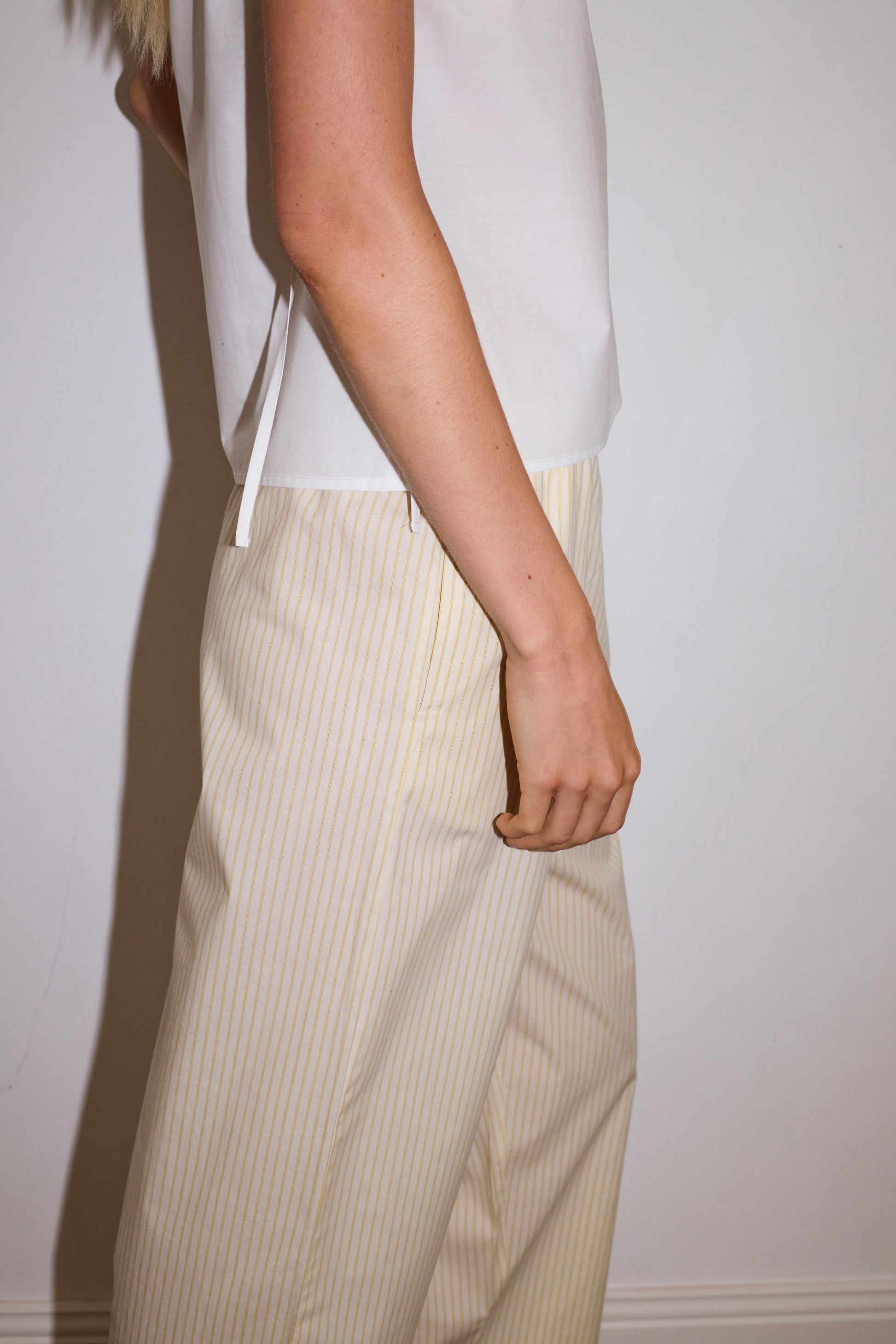 blonde female model wears a white lightweight top featuring fixed gathered armholes and right hand side armhole keyhole and ties, paired with a butter yellow striped ankle length pant features a soft minimal elastic waist with side seam pockets in a straight leg