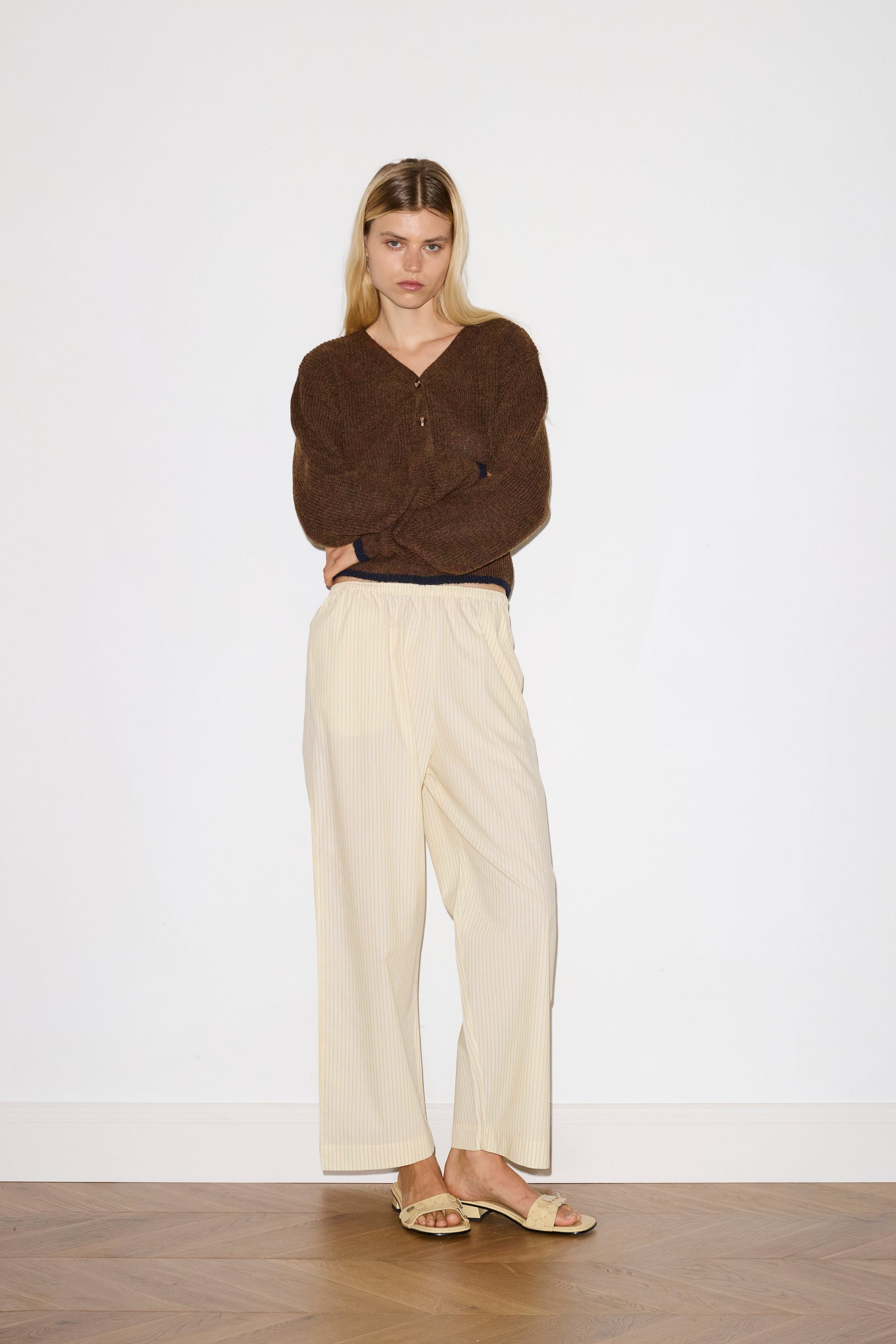 blonde female model wears long sleeve cardigan in brown merino wool fabric with navy stripe hem detail and button down front, worn with an elasticated waist yellow striped full-length trouser