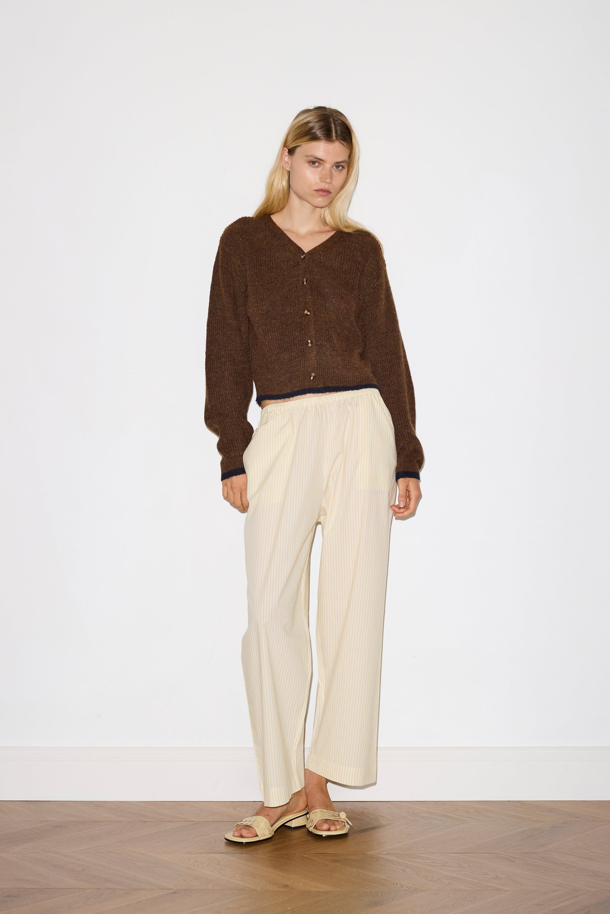 blonde female model wears long sleeve cardigan in brown merino wool fabric with navy stripe hem detail and button down front, worn with an elasticated waist yellow striped full-length trouser