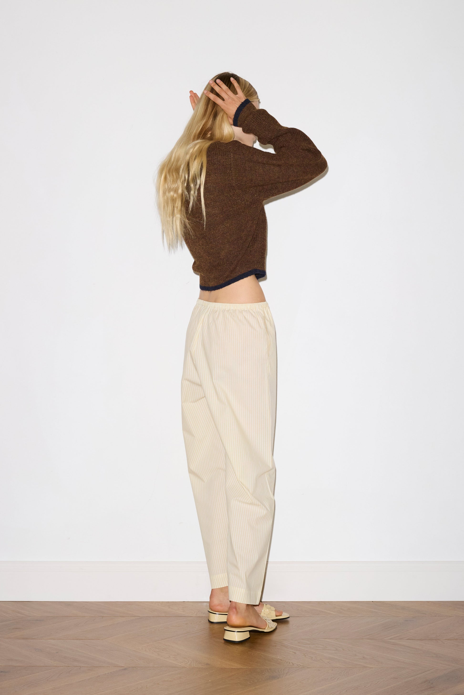 blonde female model wears long sleeve cardigan in brown merino wool fabric with navy stripe hem detail and button down front, worn with an elasticated waist yellow striped full-length trouser