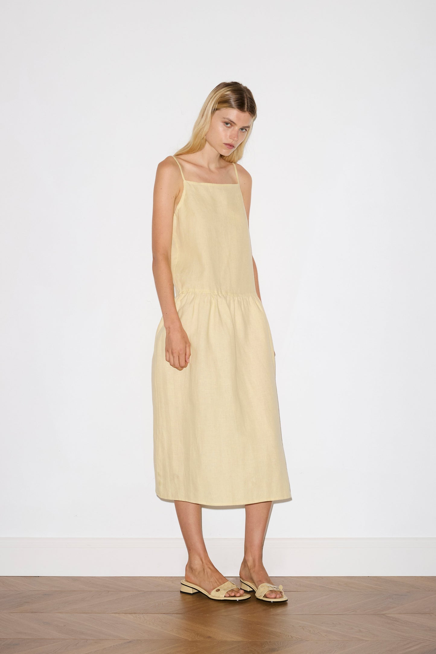 blonde haired female model wears a butter yellow relaxed mid-length dress featuring a drop waist with soft gathered skirt, gathering under the armholes and adjustable tie straps at back