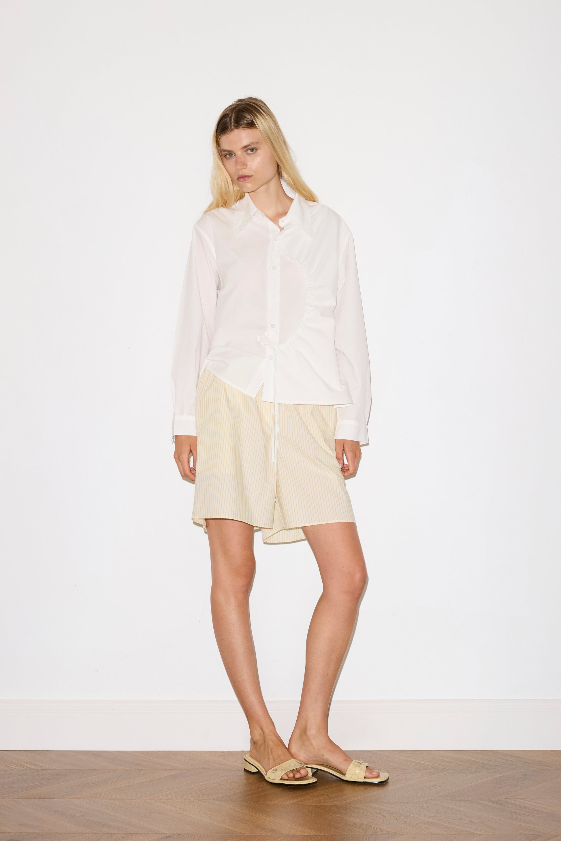 blonde female model wears a white classic crisp button up shirt in a boxy fit, featuring a dropped back yoke, dropped shoulders, a classic shirting cuff with a layered arch fabric detail on front with tie, paired with a butter yellow striped easy pull-on boxer featuring button detail at front placket, gathered elastic waistline and deep side pockets in mid length for a relaxed fit