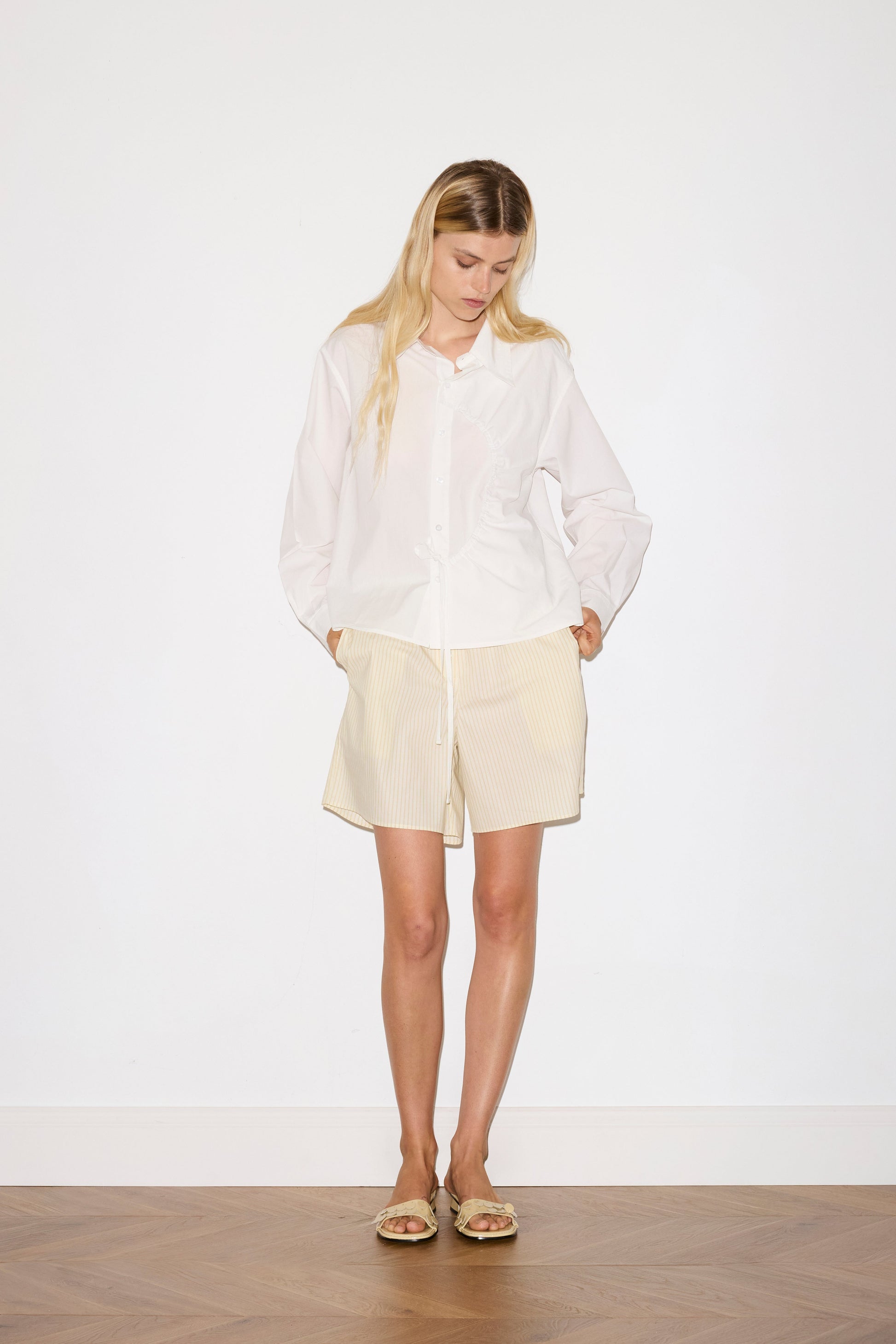 blonde female model wears a white classic crisp button up shirt in a boxy fit, featuring a dropped back yoke, dropped shoulders, a classic shirting cuff with a layered arch fabric detail on front with tie, paired with a butter yellow striped easy pull-on boxer featuring button detail at front placket, gathered elastic waistline and deep side pockets in mid length for a relaxed fit