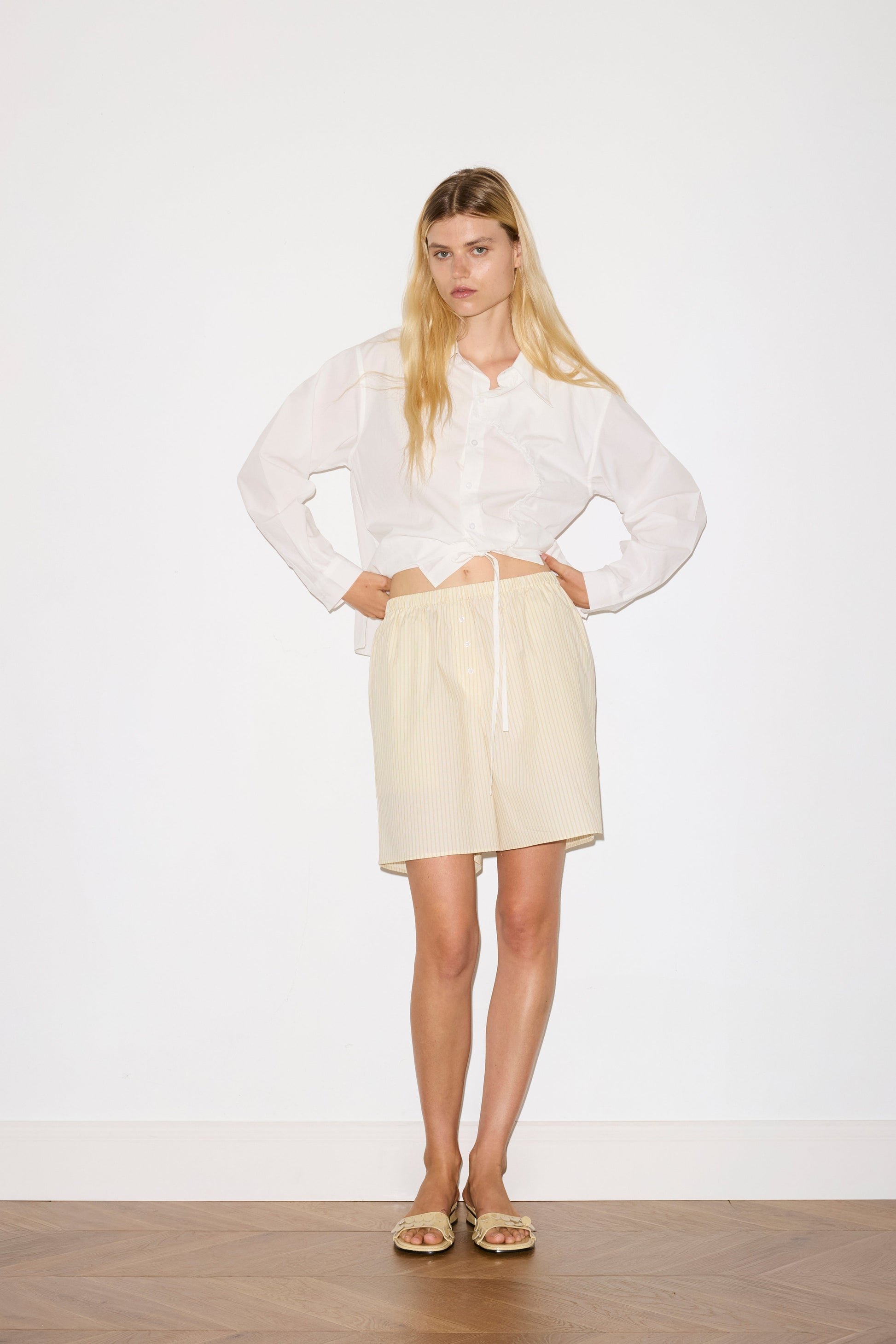 blonde female model wears a white classic crisp button up shirt in a boxy fit, featuring a dropped back yoke, dropped shoulders, a classic shirting cuff with a layered arch fabric detail on front with tie, paired with a butter yellow striped easy pull-on boxer featuring button detail at front placket, gathered elastic waistline and deep side pockets in mid length for a relaxed fit