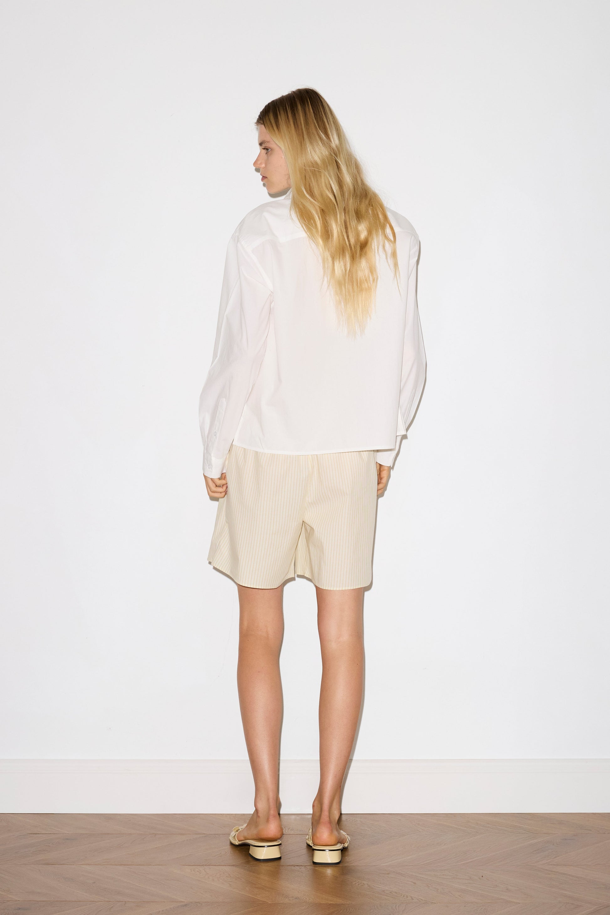 blonde female model wears a white classic crisp button up shirt in a boxy fit, featuring a dropped back yoke, dropped shoulders, a classic shirting cuff with a layered arch fabric detail on front with tie, paired with a butter yellow striped easy pull-on boxer featuring button detail at front placket, gathered elastic waistline and deep side pockets in mid length for a relaxed fit