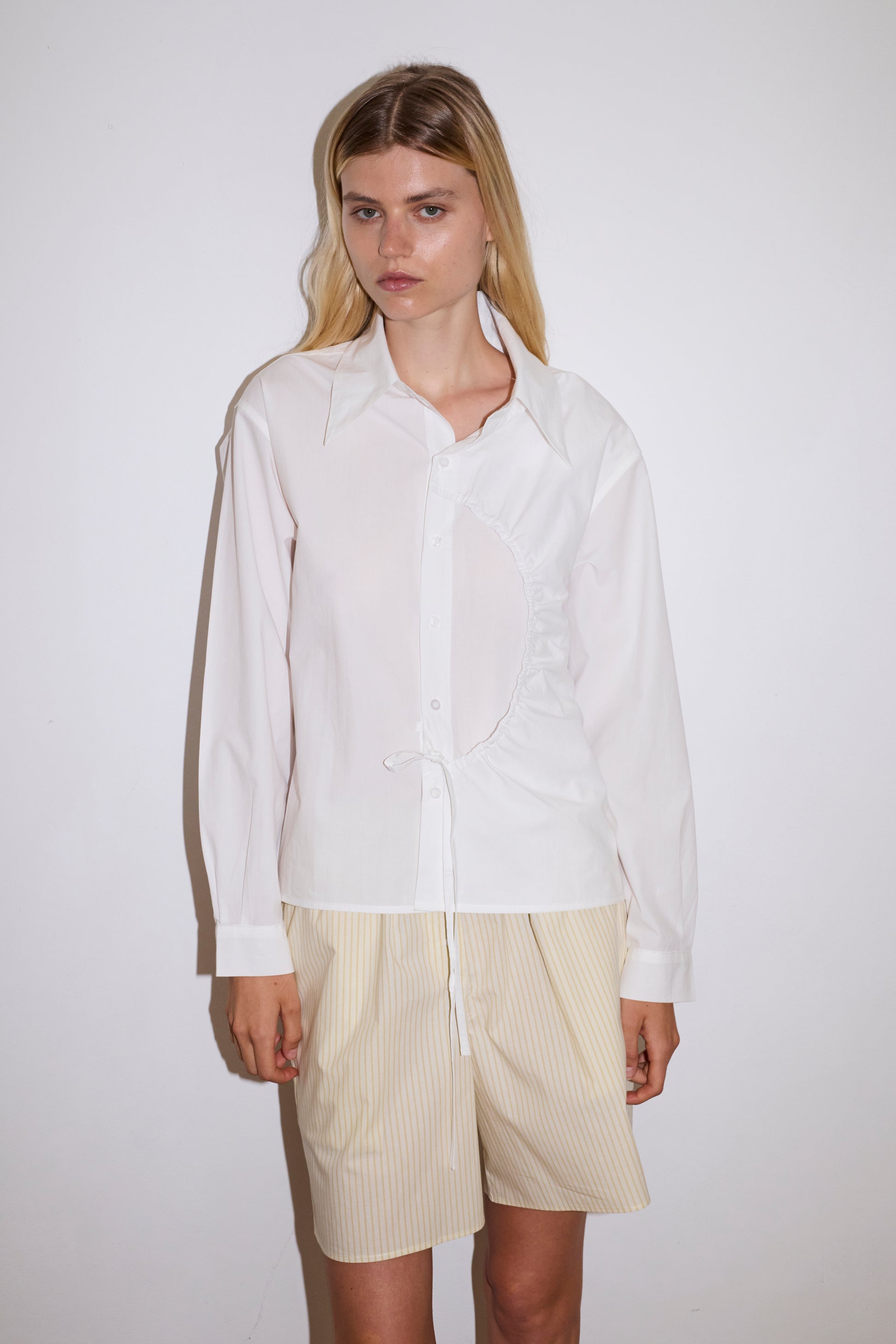 blonde female model wears a white classic crisp button up shirt in a boxy fit, featuring a dropped back yoke, dropped shoulders, a classic shirting cuff with a layered arch fabric detail on front with tie, paired with a butter yellow striped easy pull-on boxer featuring button detail at front placket, gathered elastic waistline and deep side pockets in mid length for a relaxed fit