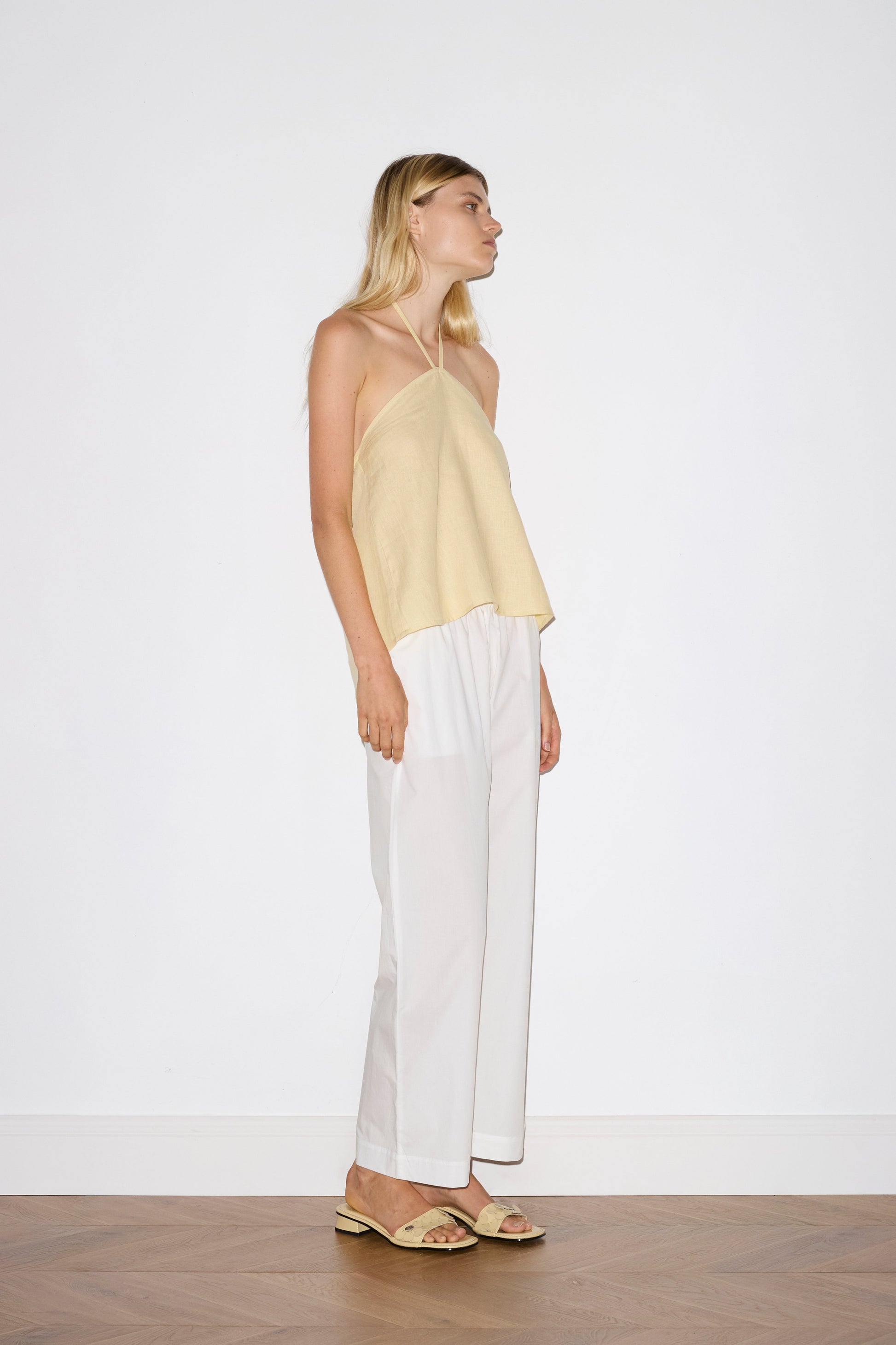 blonde haired female model wears a yellow relaxed halter top features flattering straight neckline, fine tie halter straps and snap closure at back with button detail, worn with a white  ankle length pant features a soft minimal elastic waist with side seam pockets in a straight leg. 