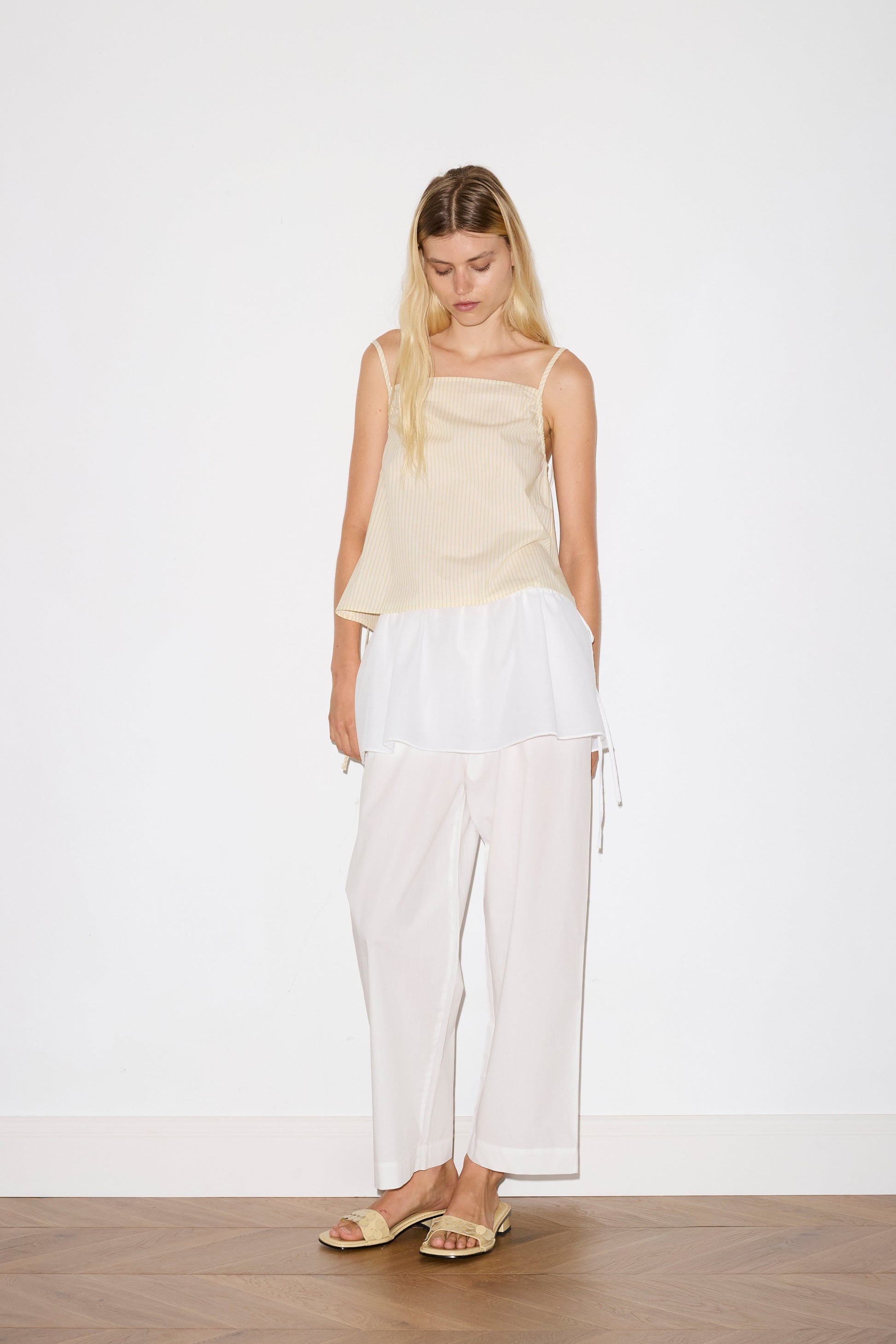 blonde female model wears a white lightweight wrap belt features a drawcord casing, adjustable ties to wrap the body, and a babylock hem, paired with a white cotton trouser and butter striped yellow lightweight top featuring fixed gathered armholes and right hand side armhole keyhole and ties