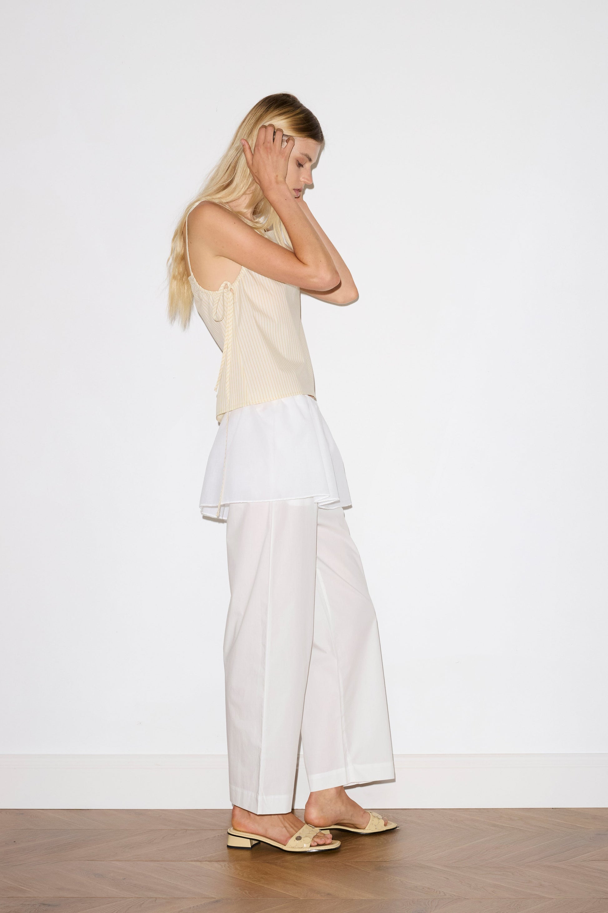 blonde female model wears a white lightweight wrap belt features a drawcord casing, adjustable ties to wrap the body, and a babylock hem, paired with a white cotton trouser and butter striped yellow lightweight top featuring fixed gathered armholes and right hand side armhole keyhole and ties