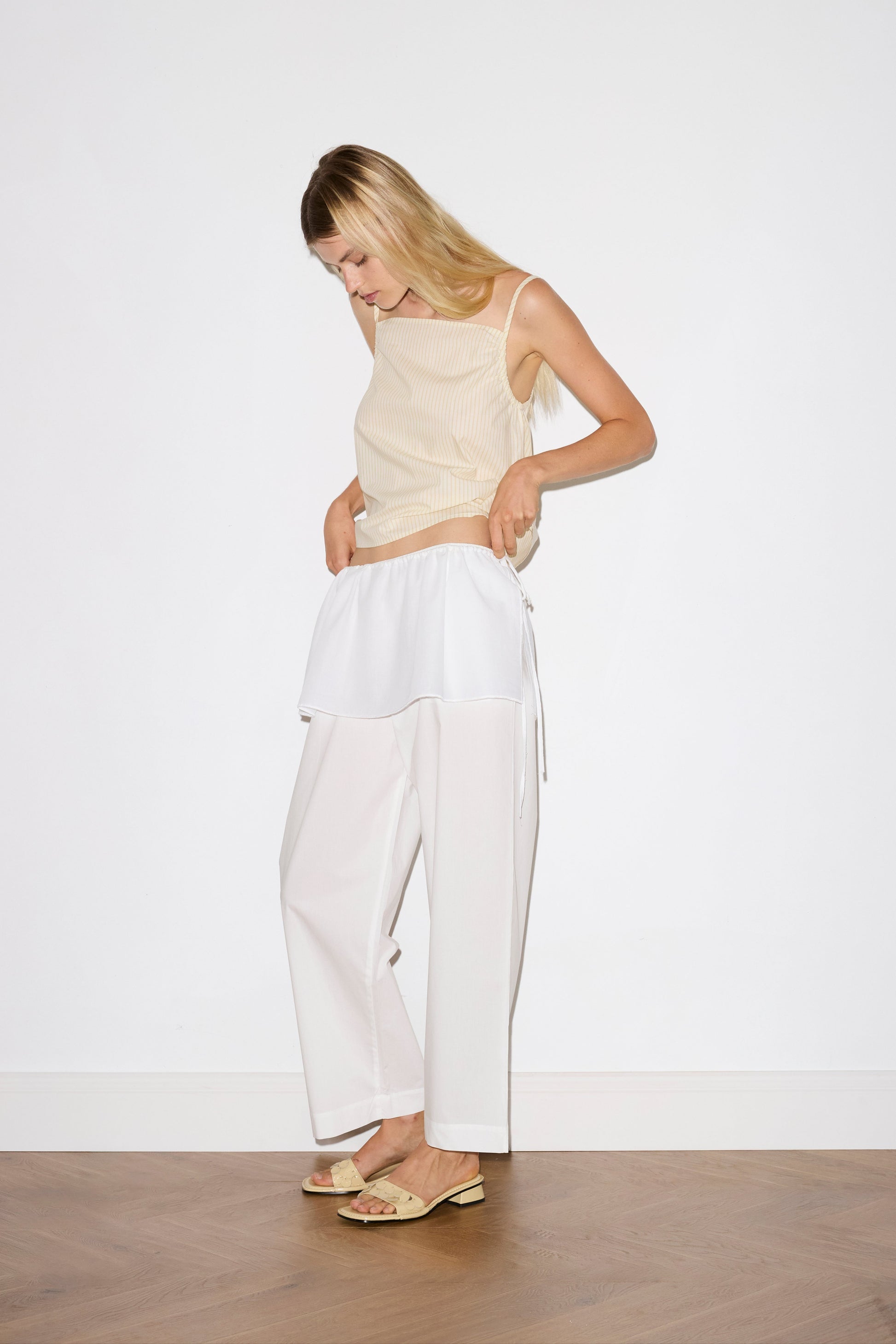 blonde female model wears a white lightweight wrap belt features a drawcord casing, adjustable ties to wrap the body, and a babylock hem, paired with a white cotton trouser and butter striped yellow lightweight top featuring fixed gathered armholes and right hand side armhole keyhole and ties