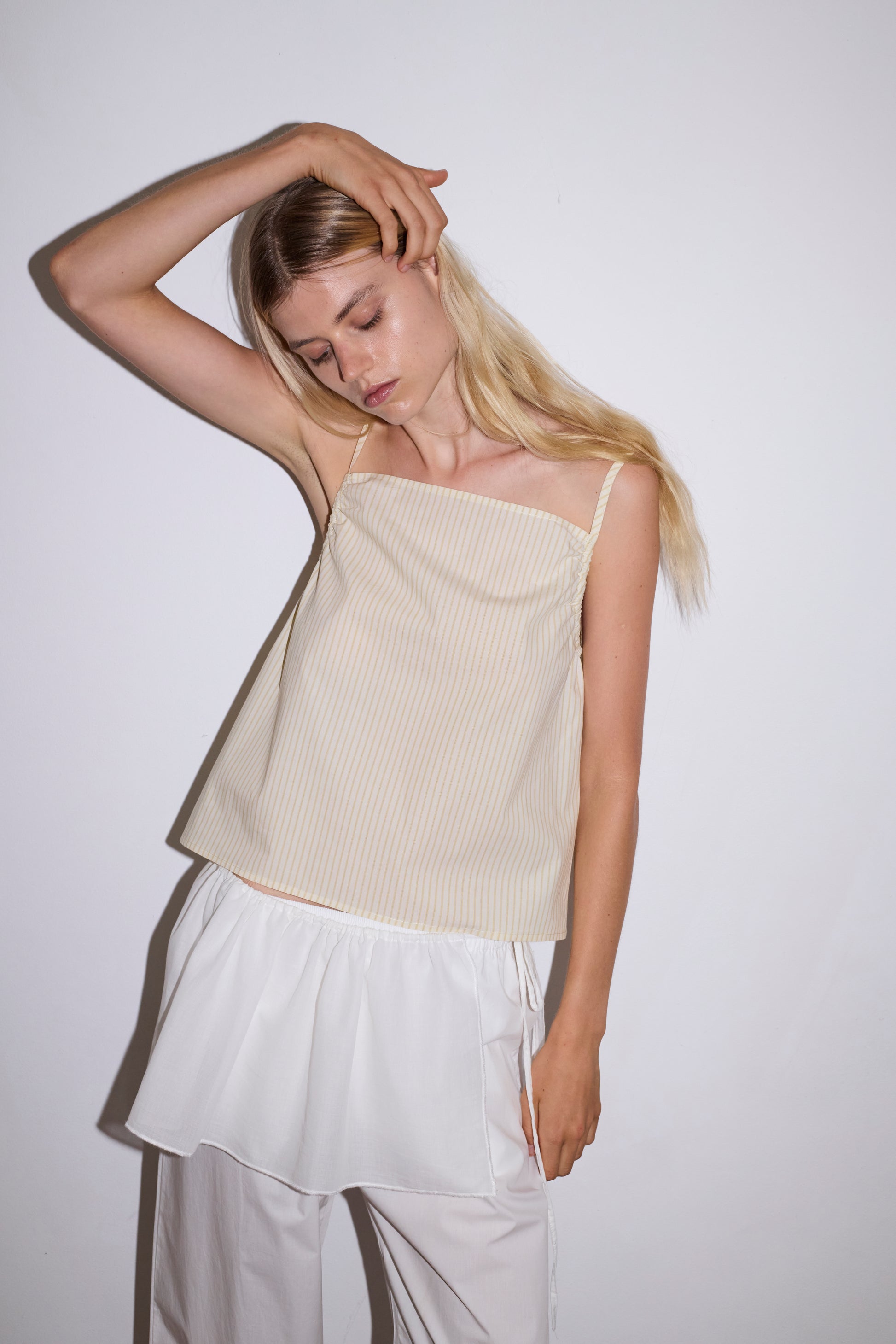 blonde female model wears a white lightweight wrap belt features a drawcord casing, adjustable ties to wrap the body, and a babylock hem, paired with a white cotton trouser and butter striped yellow lightweight top featuring fixed gathered armholes and right hand side armhole keyhole and ties