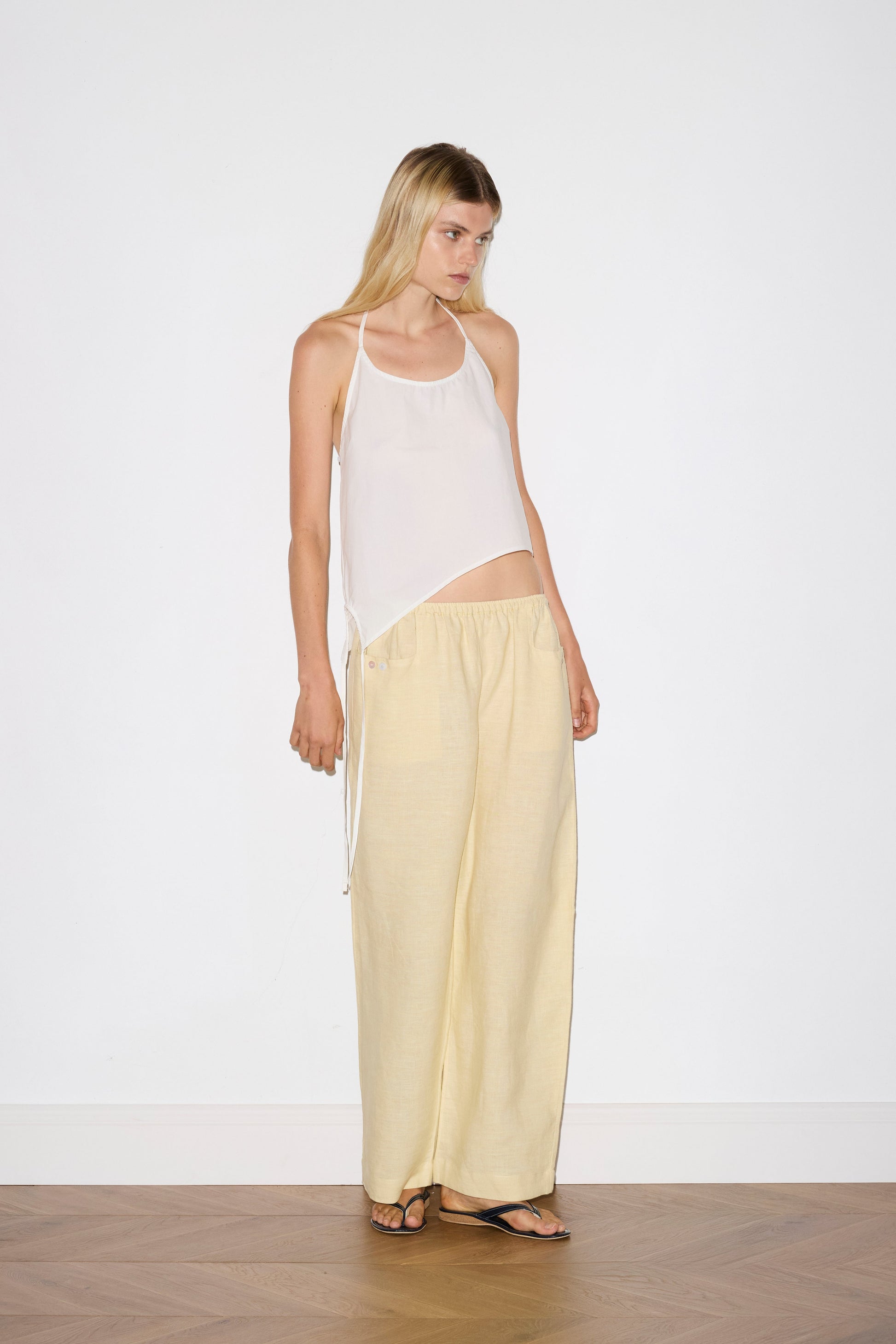 blonde haired female model wears a butter yellow ankle length, straight leg pant features a soft minimal elastic waist with front seam pockets and button detail, paired with a white relaxed halter top features flattering curved neckline, asymmetrical bottom hem feature with side tie opening, keyhole with button closure at side and fine tie halter straps.