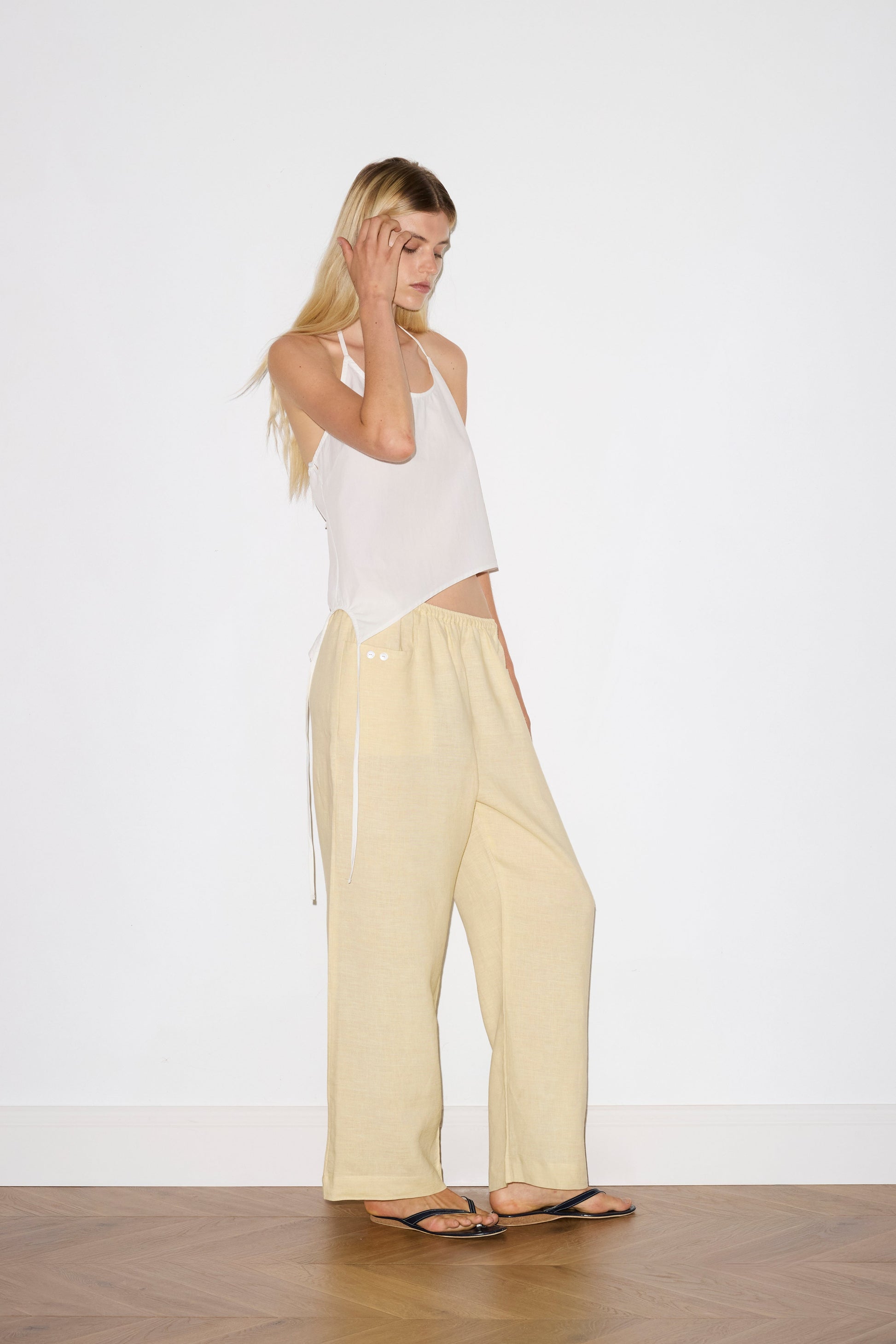 blonde haired female model wears a butter yellow ankle length, straight leg pant features a soft minimal elastic waist with front seam pockets and button detail, paired with a white relaxed halter top features flattering curved neckline, asymmetrical bottom hem feature with side tie opening, keyhole with button closure at side and fine tie halter straps.