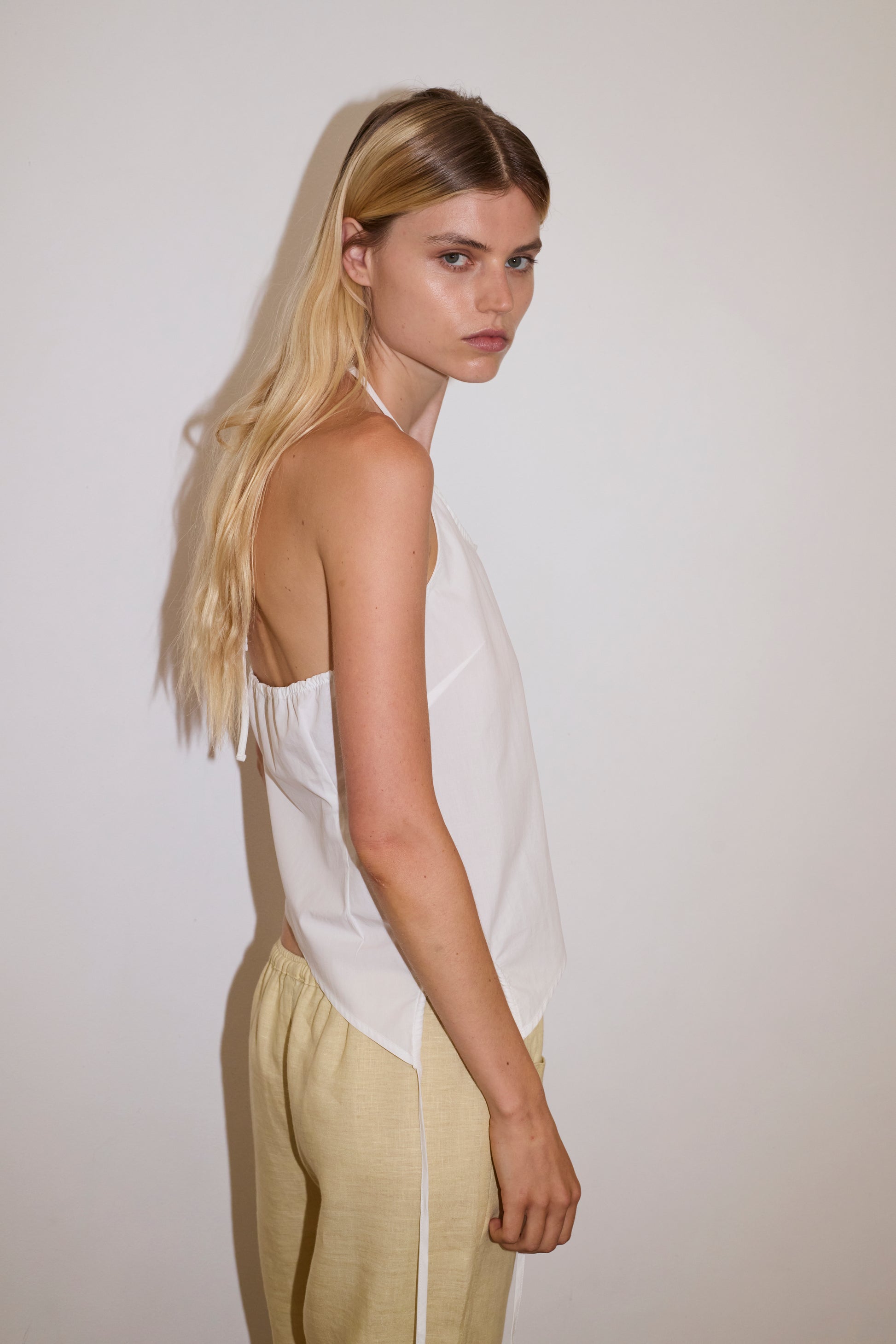 blonde haired female model wears a butter yellow ankle length, straight leg pant features a soft minimal elastic waist with front seam pockets and button detail, paired with a white relaxed halter top features flattering curved neckline, asymmetrical bottom hem feature with side tie opening, keyhole with button closure at side and fine tie halter straps.