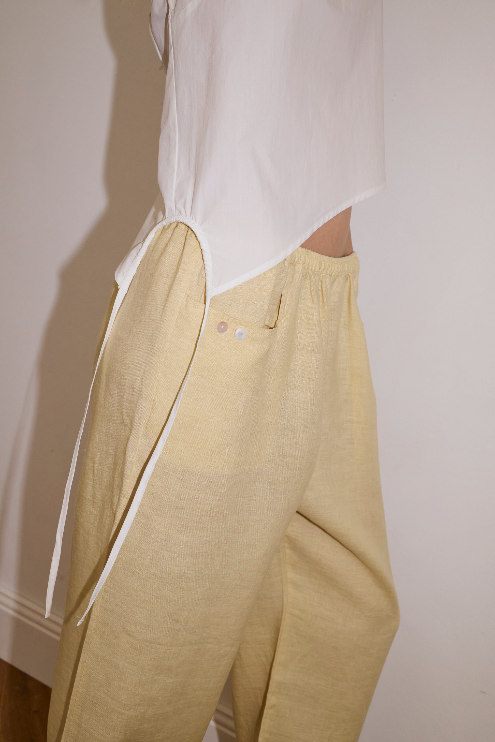 blonde haired female model wears a butter yellow ankle length, straight leg pant features a soft minimal elastic waist with front seam pockets and button detail, paired with a white relaxed halter top features flattering curved neckline, asymmetrical bottom hem feature with side tie opening, keyhole with button closure at side and fine tie halter straps.