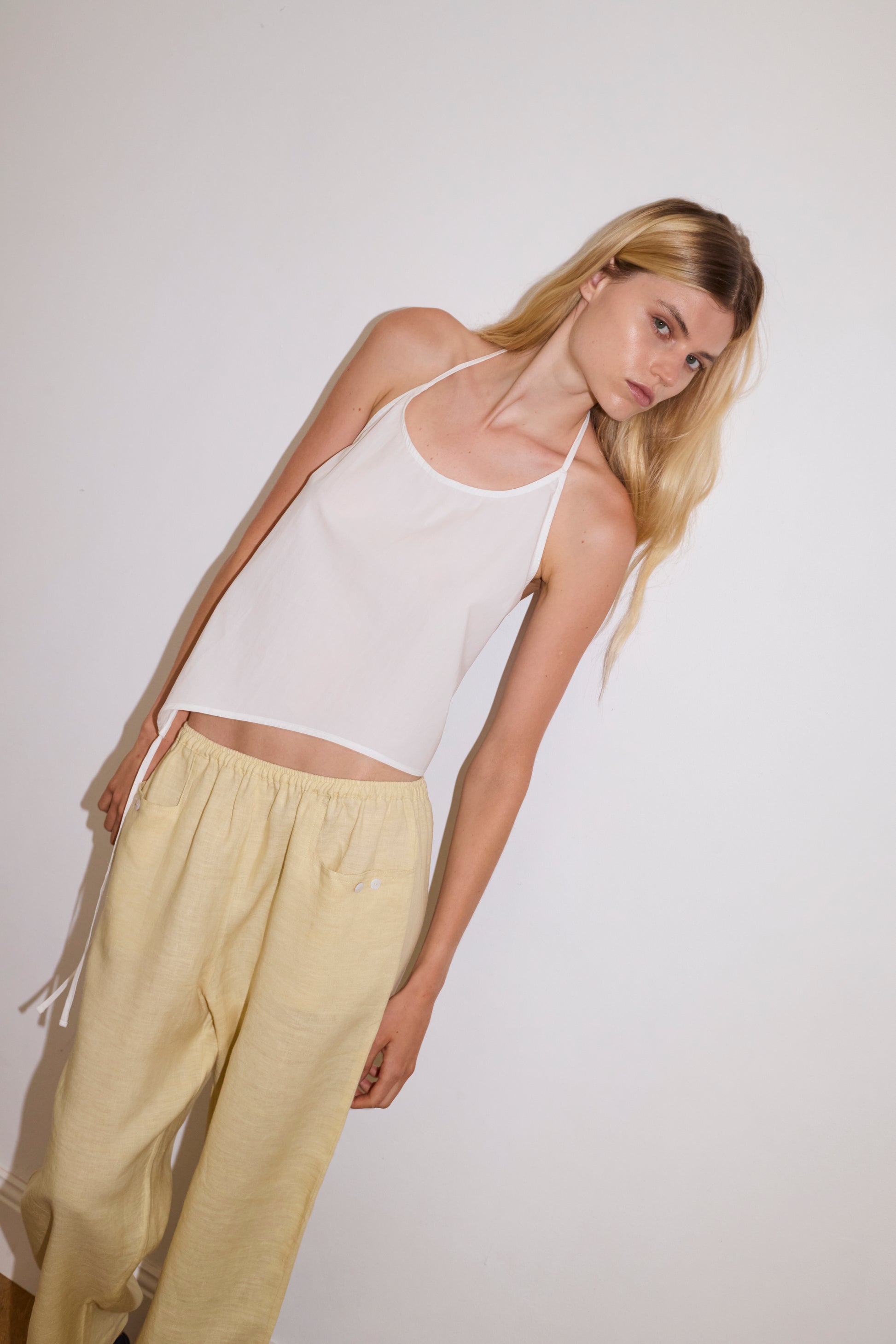 blonde haired female model wears a butter yellow ankle length, straight leg pant features a soft minimal elastic waist with front seam pockets and button detail, paired with a white relaxed halter top features flattering curved neckline, asymmetrical bottom hem feature with side tie opening, keyhole with button closure at side and fine tie halter straps.