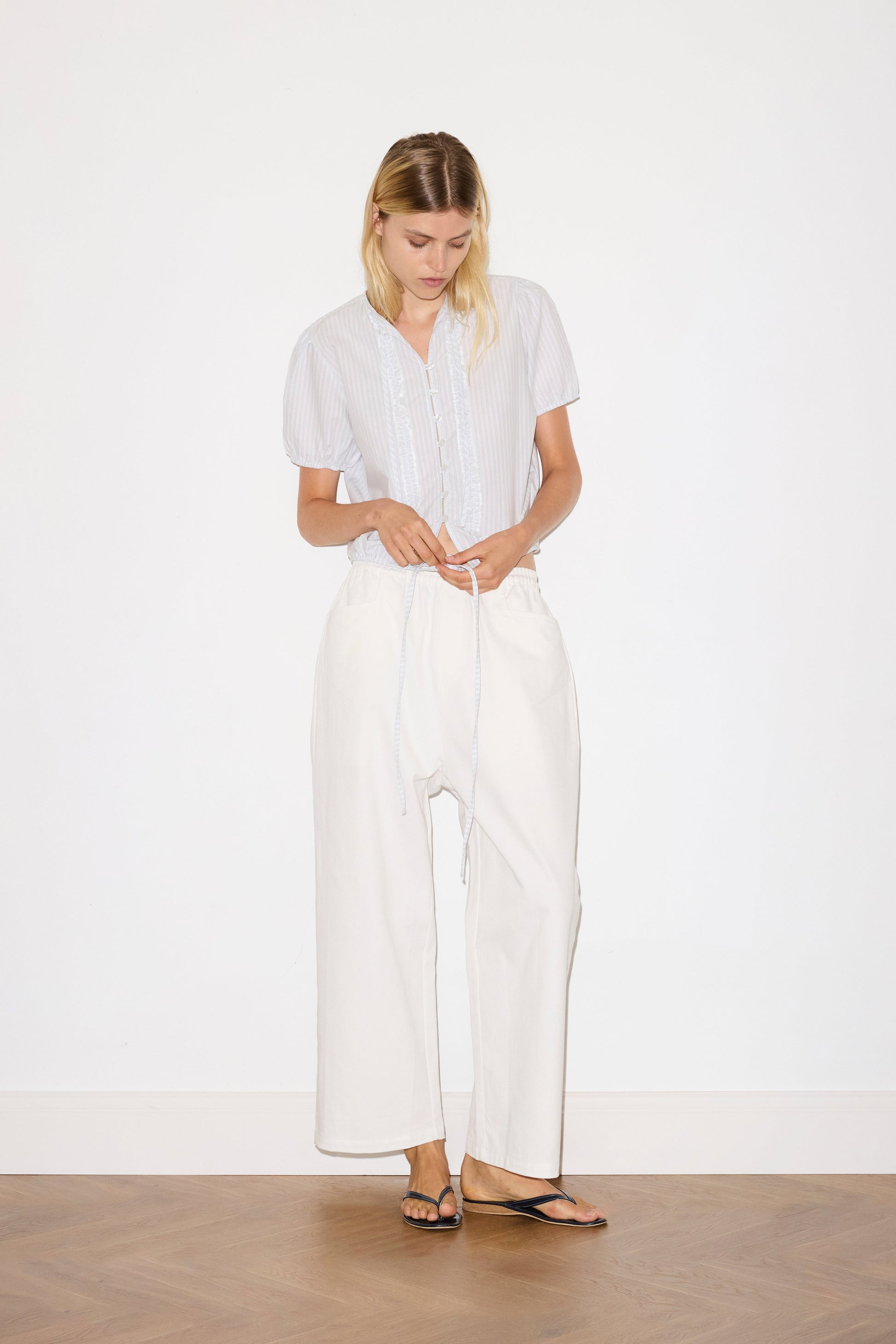 blonde female model wears a white Heavy- weighted relaxed-fit pant featuring a wide-leg silhouette, elasticated waistband, and square pocket detail, paired with a sage and white striped elevated everyday top features a flattering v neckline with button down front, fixed ruffle details, soft bloussant sleeve and gathered elastic cuffs