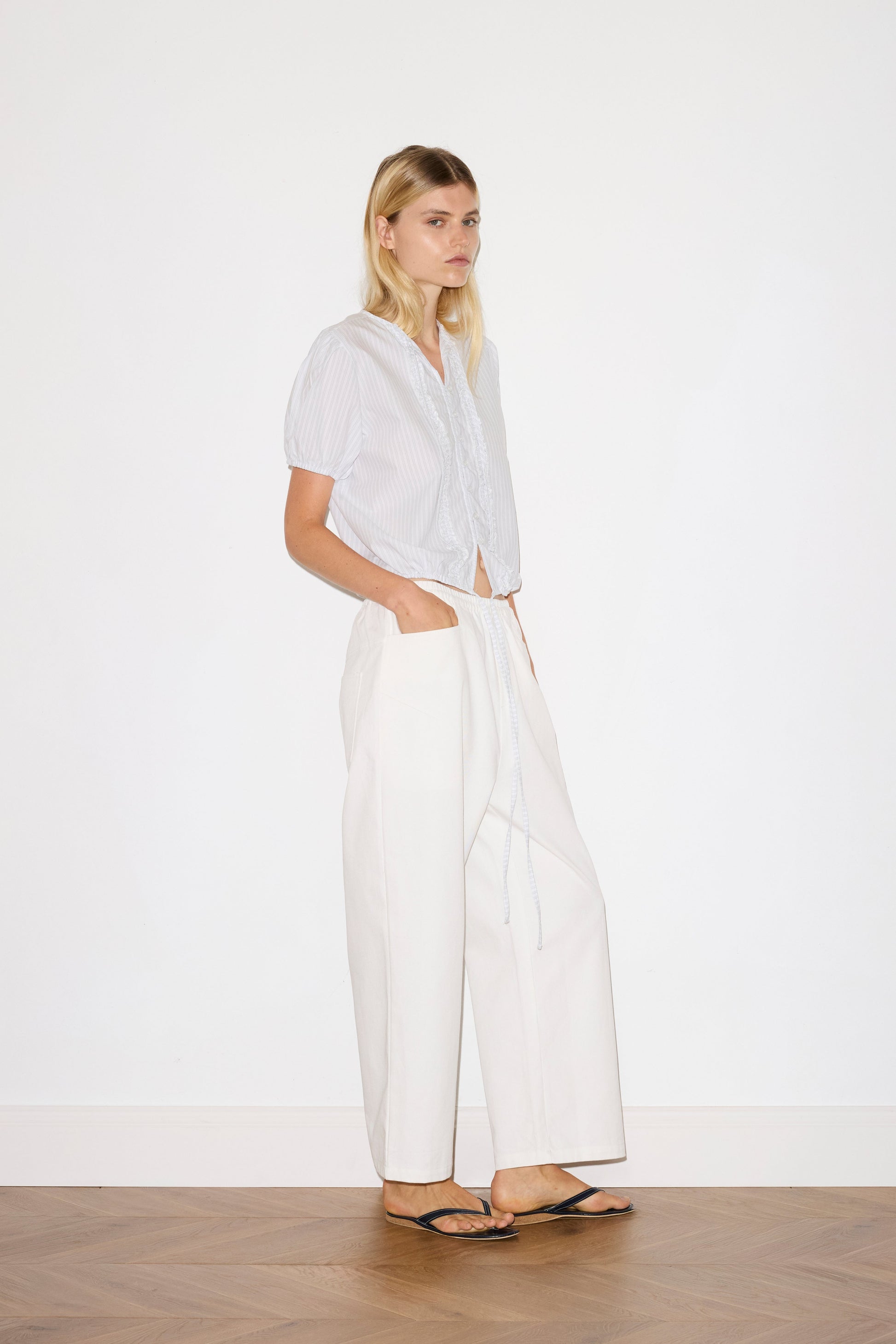 blonde haired female model wears a sage and white striped elevated everyday top features a flattering v neckline with button down front, fixed ruffle details, soft bloussant sleeve and gathered elastic cuffs, paired with a white Heavy- weighted relaxed-fit pant featuring a wide-leg silhouette, elasticated waistband, and square pocket detail