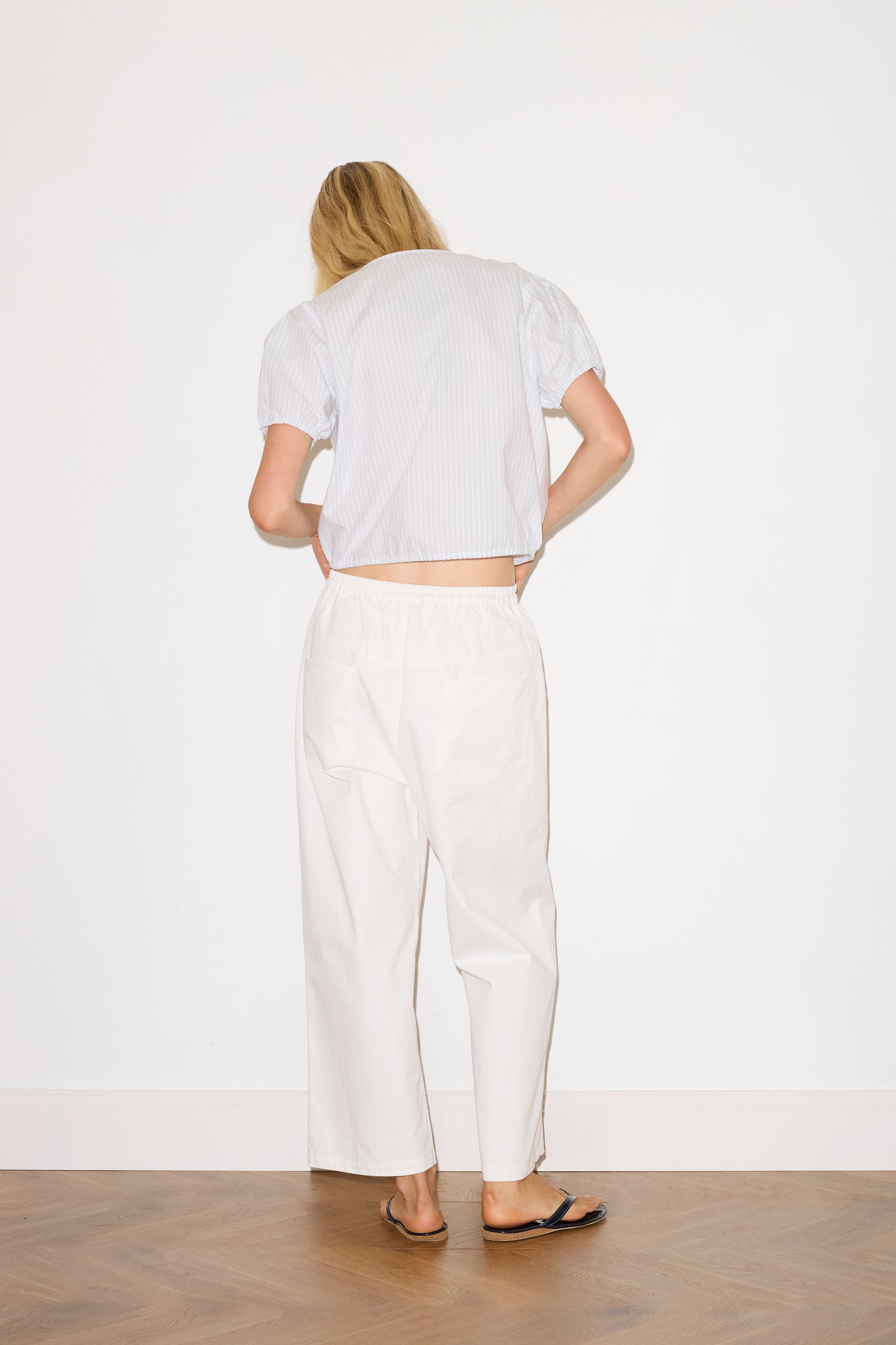 blonde female model wears a white Heavy- weighted relaxed-fit pant featuring a wide-leg silhouette, elasticated waistband, and square pocket detail, paired with a sage and white striped elevated everyday top features a flattering v neckline with button down front, fixed ruffle details, soft bloussant sleeve and gathered elastic cuffs