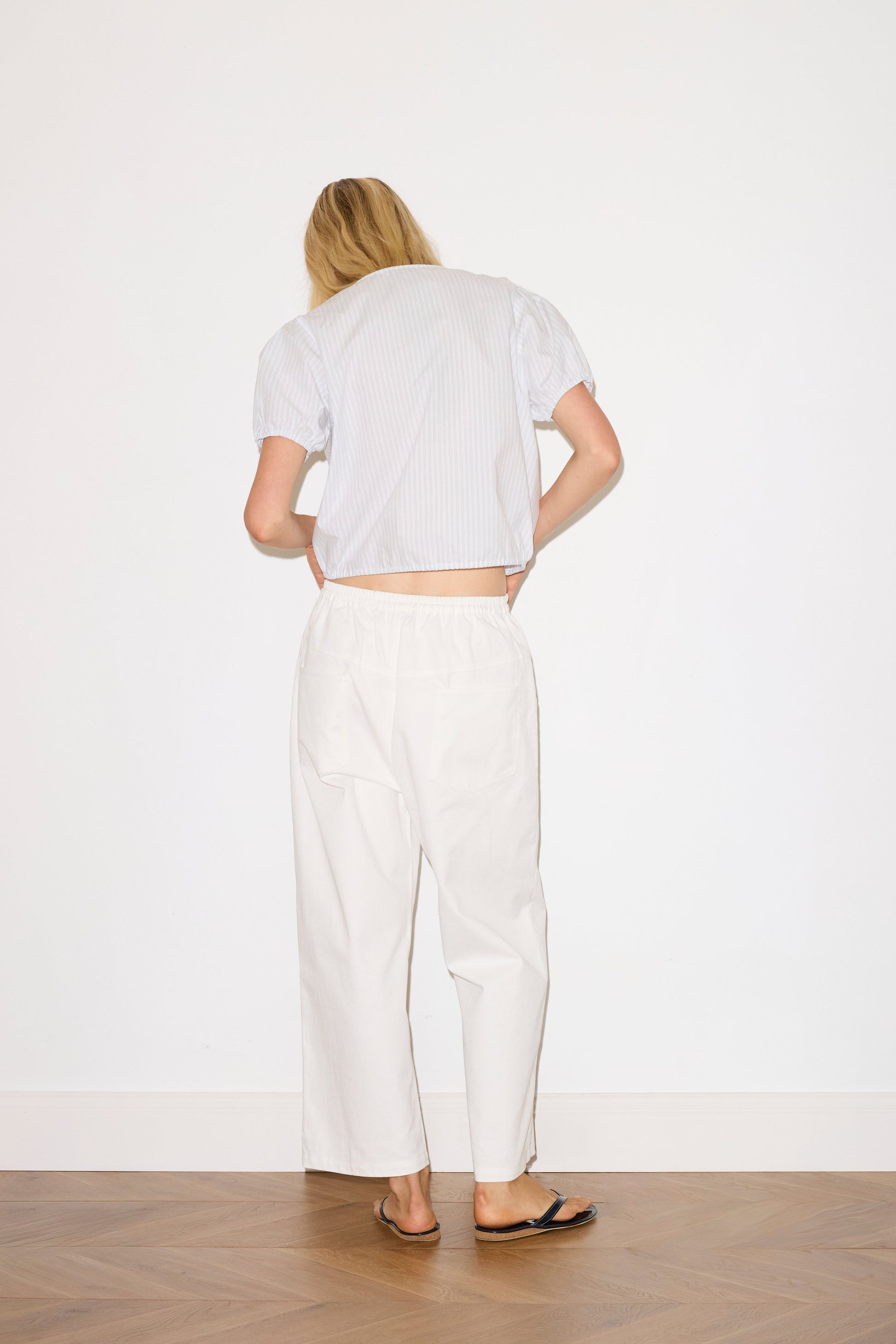 blonde haired female model wears a sage and white striped elevated everyday top features a flattering v neckline with button down front, fixed ruffle details, soft bloussant sleeve and gathered elastic cuffs, paired with a white Heavy- weighted relaxed-fit pant featuring a wide-leg silhouette, elasticated waistband, and square pocket detail