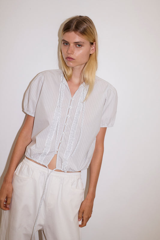 blonde haired female model wears a sage and white striped elevated everyday top features a flattering v neckline with button down front, fixed ruffle details, soft bloussant sleeve and gathered elastic cuffs, paired with a white Heavy- weighted relaxed-fit pant featuring a wide-leg silhouette, elasticated waistband, and square pocket detail