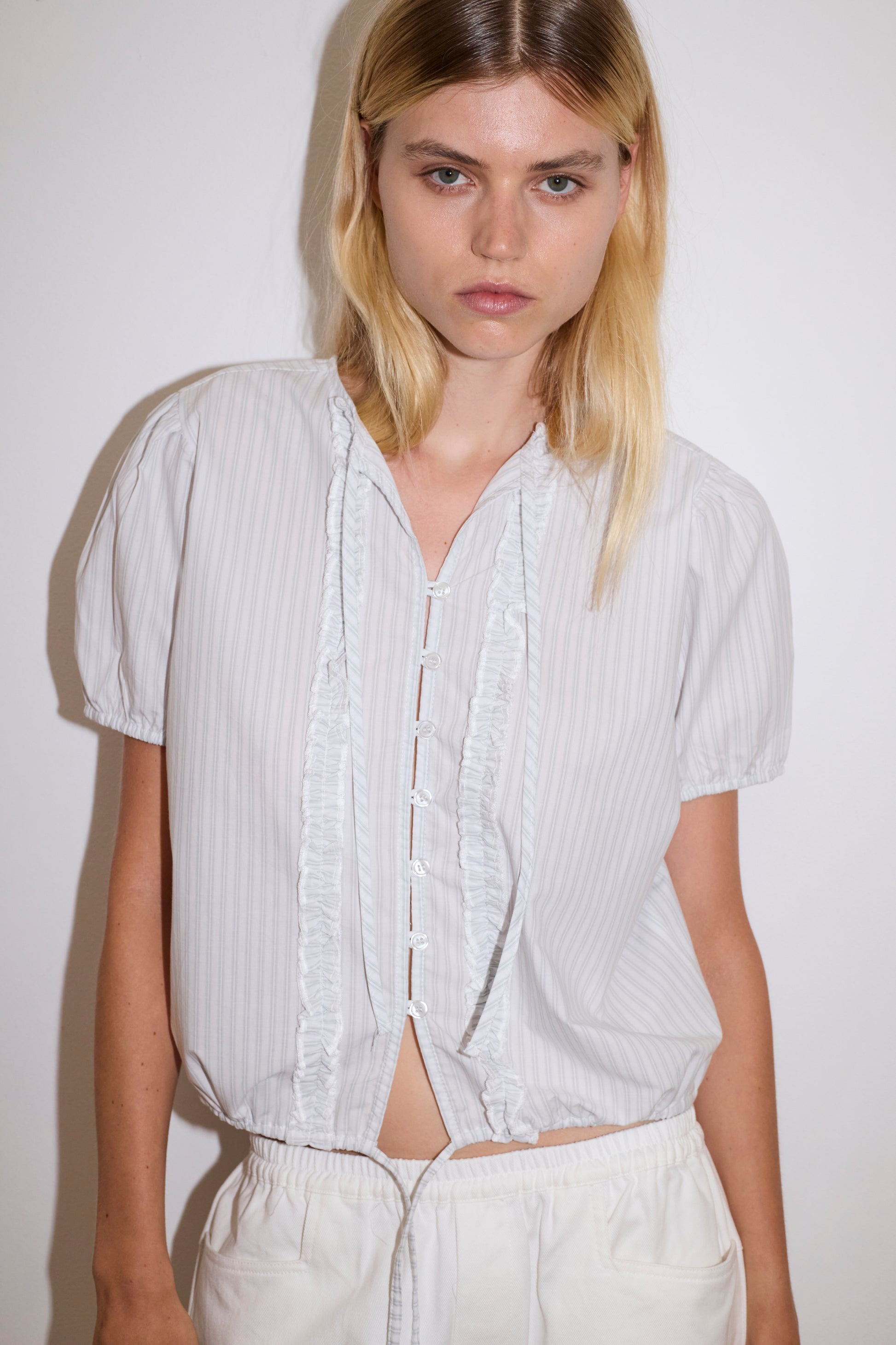 blonde haired female model wears a sage and white striped elevated everyday top features a flattering v neckline with button down front, fixed ruffle details, soft bloussant sleeve and gathered elastic cuffs, paired with a white Heavy- weighted relaxed-fit pant featuring a wide-leg silhouette, elasticated waistband, and square pocket detail