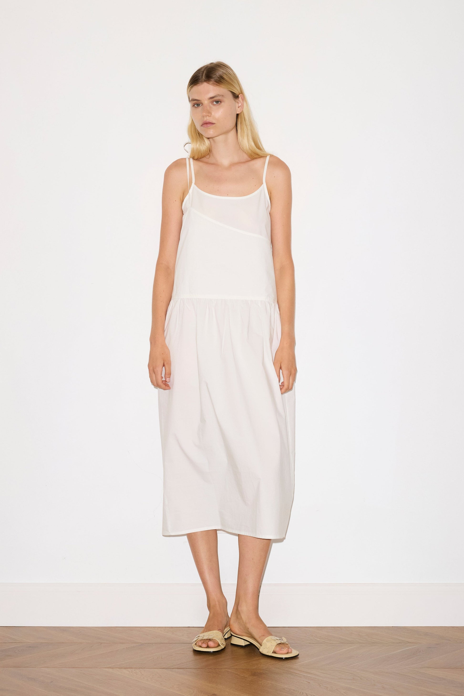 blonde female model wears a white mid-length dress featuring adjustable underlayer straps with sliders for a customised fit and a tied overlayer strap detail at the shoulder