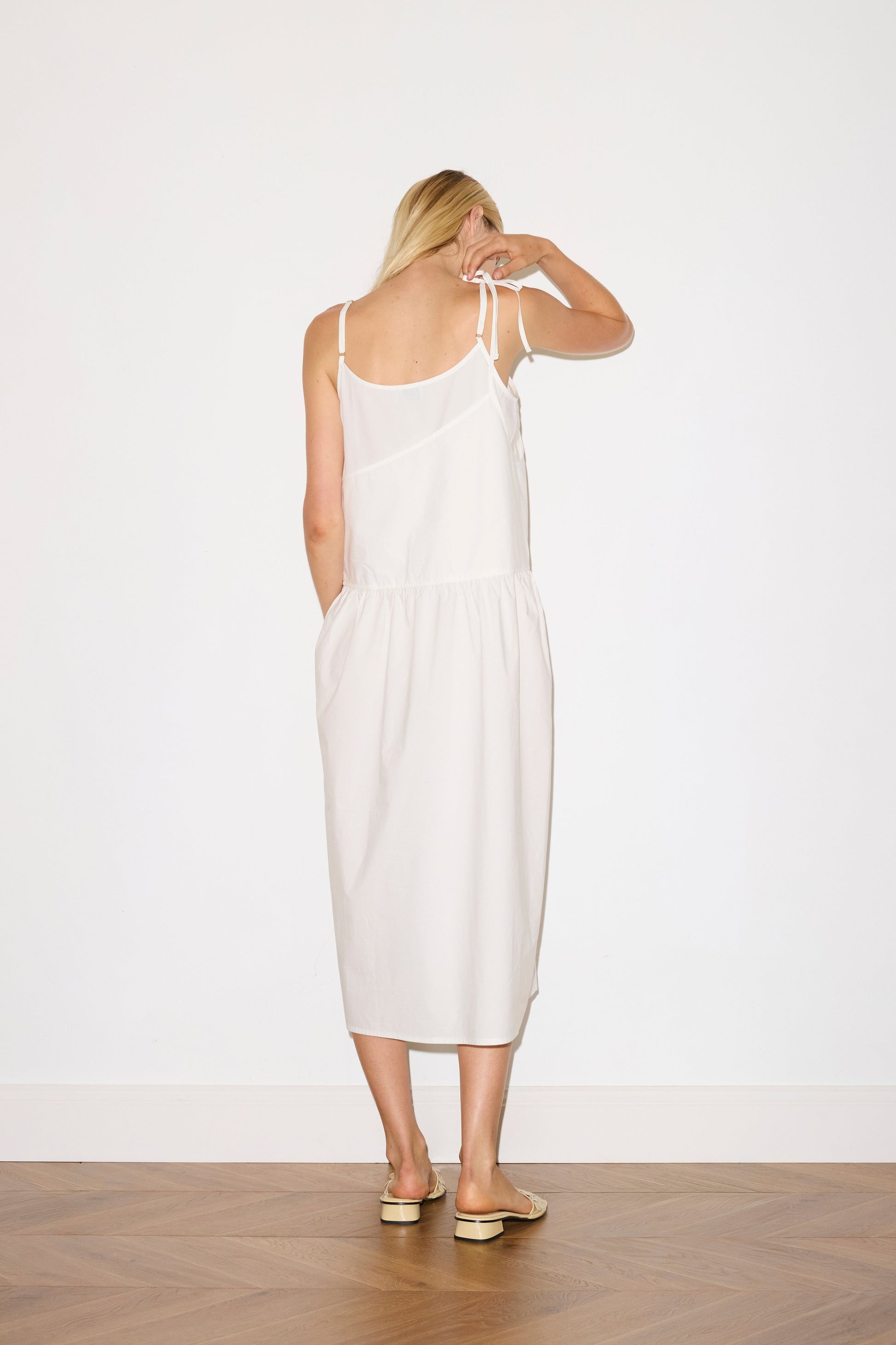 blonde female model wears a white mid-length dress featuring adjustable underlayer straps with sliders for a customised fit and a tied overlayer strap detail at the shoulder