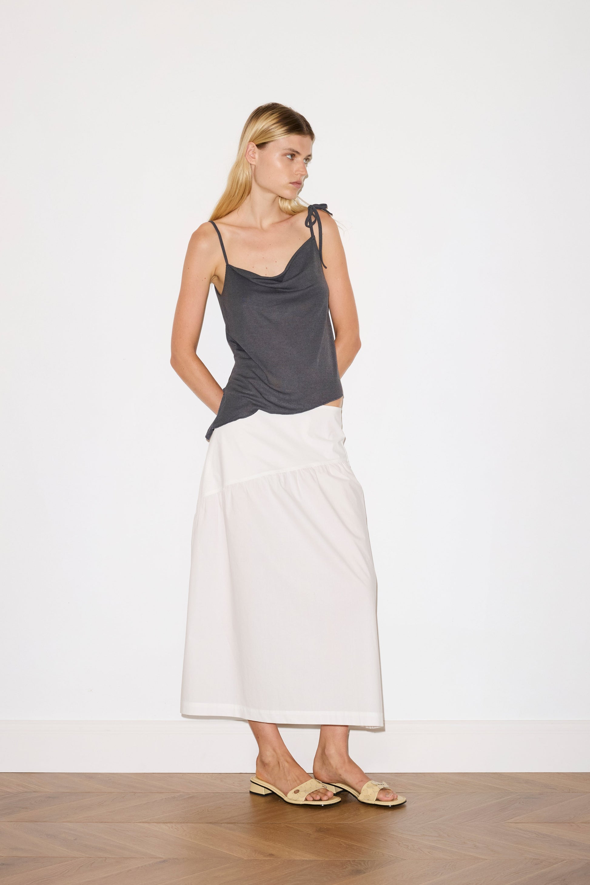 blonde female model wears a sheer fine knit relaxed tank with a fold over cowl neckline and low draping back, features self fabric rouleau straps, baby lock hem finishes and a asymmetric bottom detail, worn with a midi length white skirt 