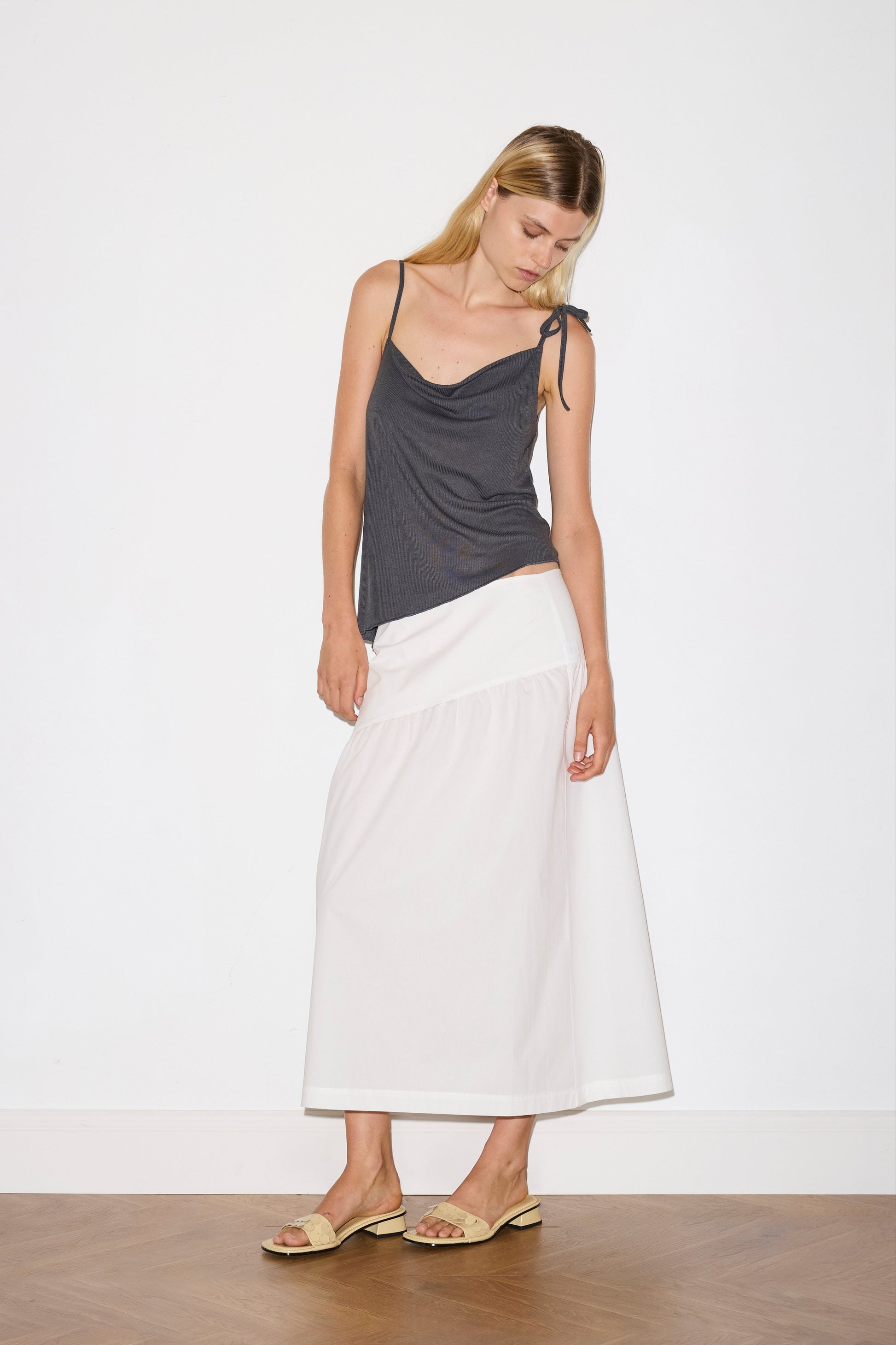 blonde female model wears a sheer fine knit relaxed tank with a fold over cowl neckline and low draping back, features self fabric rouleau straps, baby lock hem finishes and a asymmetric bottom detail, worn with a midi length white skirt 