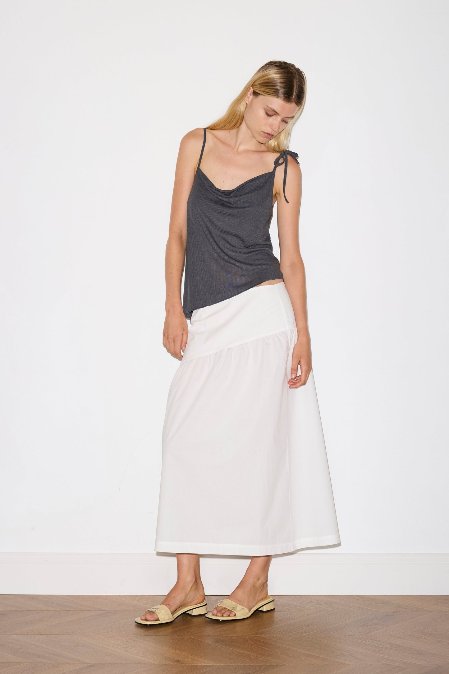 blonde female model wears a sheer fine knit relaxed tank with a fold over cowl neckline and low draping back, features self fabric rouleau straps, baby lock hem finishes and a asymmetric bottom detail, worn with a midi length white skirt 