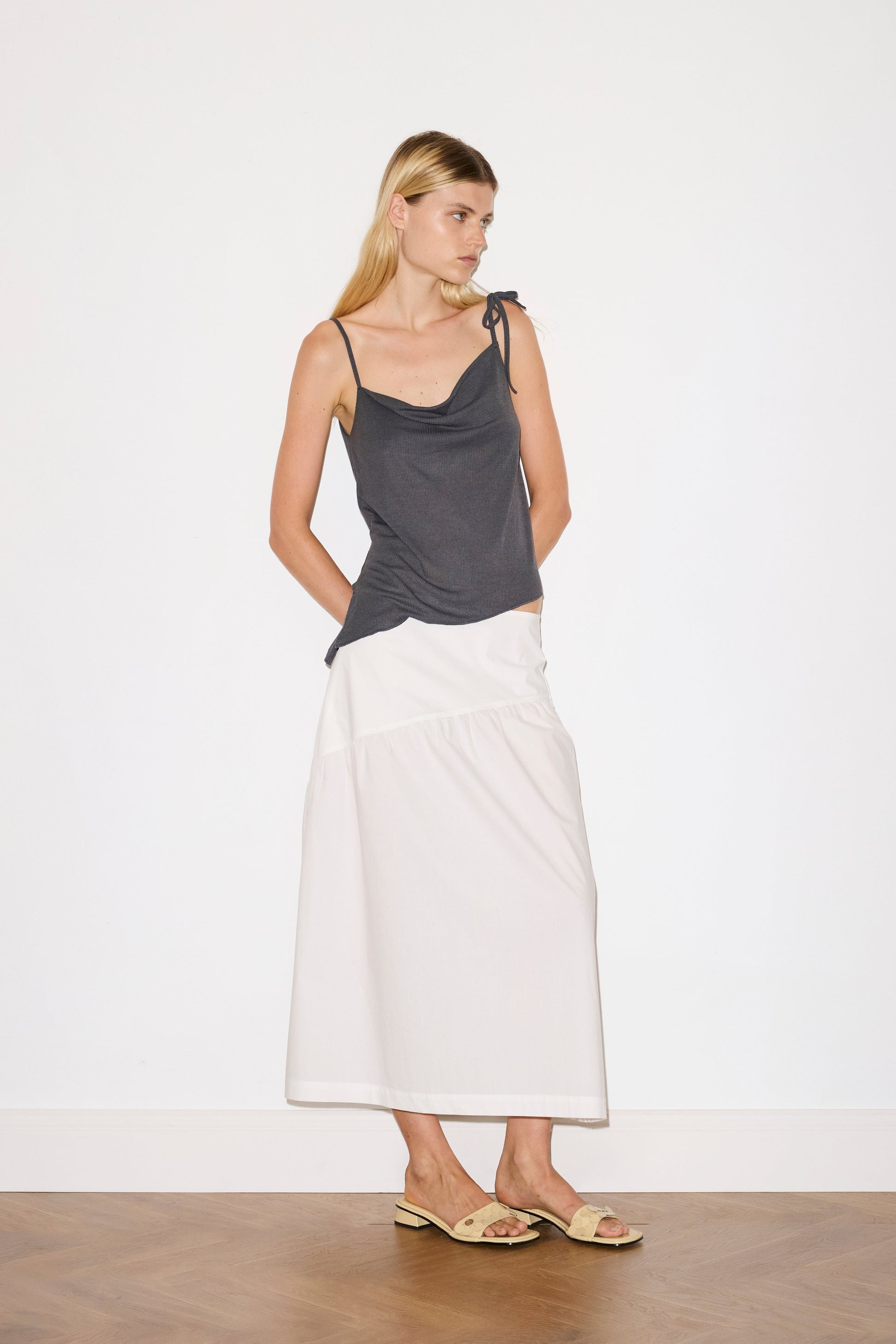 blonde female model wears a sheer fine knit relaxed tank with a fold over cowl neckline and low draping back, features self fabric rouleau straps, baby lock hem finishes and a asymmetric bottom detail, worn with a midi length white skirt 