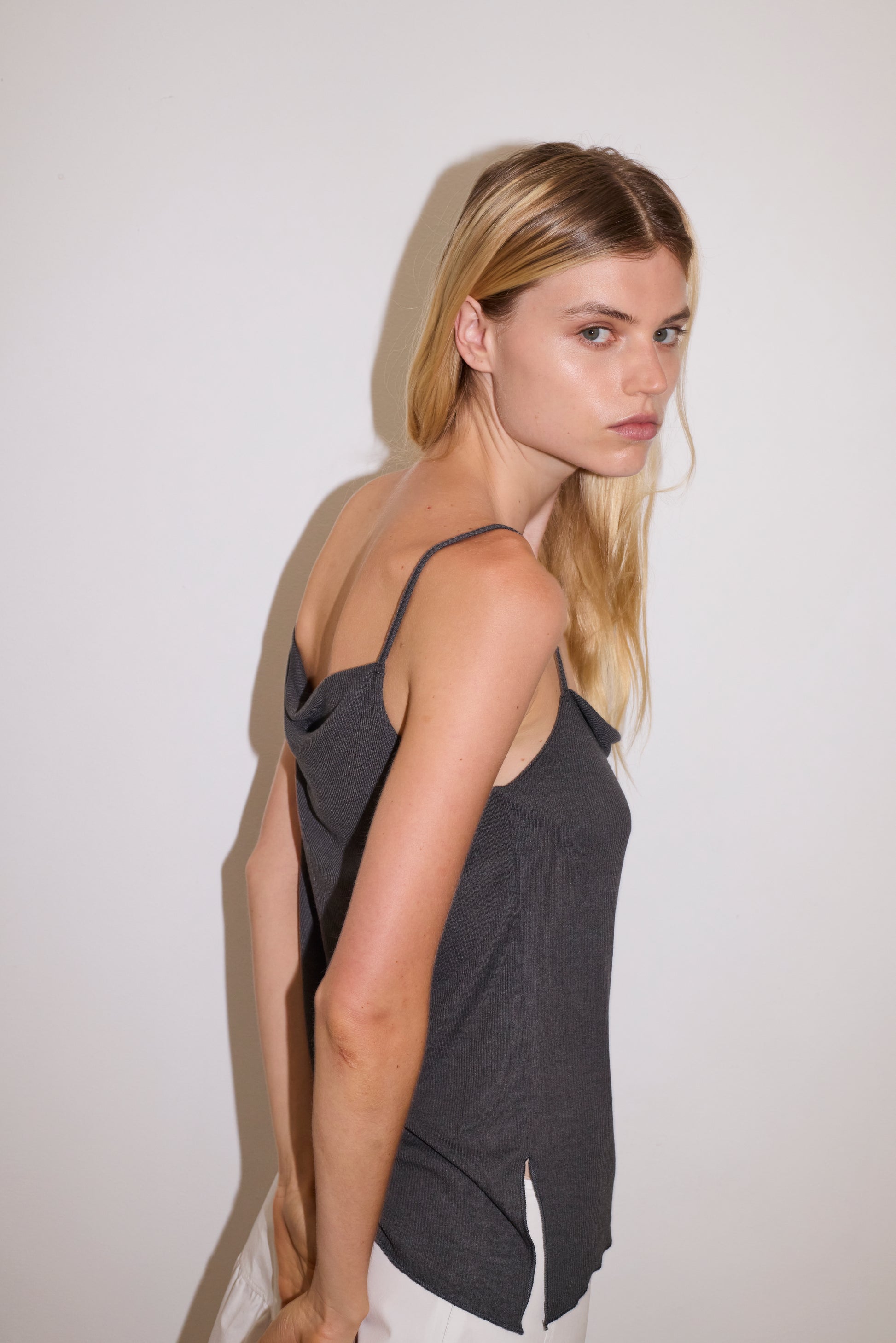 blonde female model wears a sheer fine knit relaxed tank with a fold over cowl neckline and low draping back, features self fabric rouleau straps, baby lock hem finishes and a asymmetric bottom detail, worn with a midi length white skirt 