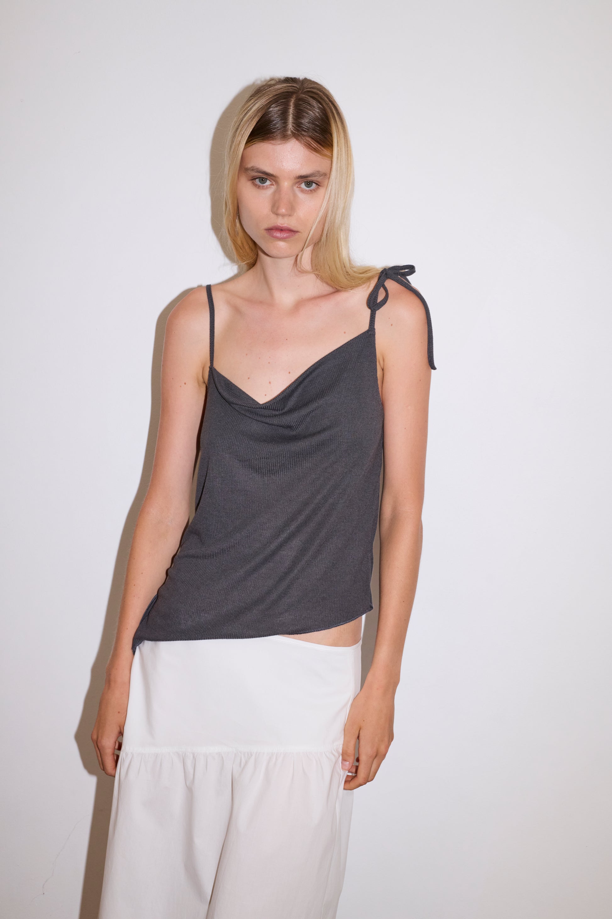 blonde female model wears a sheer fine knit relaxed tank with a fold over cowl neckline and low draping back, features self fabric rouleau straps, baby lock hem finishes and a asymmetric bottom detail, worn with a midi length white skirt 