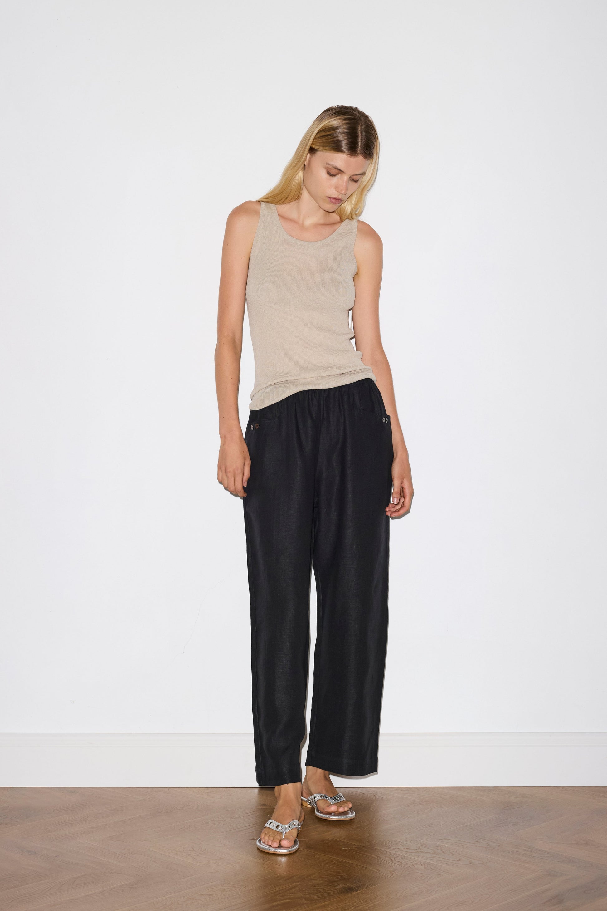 blonde female model wears a beige minimalist tank-style top featuring raw edge detailing and an asymmetrical tie strap design for a unique and adjustable fit. made with a lightweight ribbed texture, worn with a black ankle length, straight leg pant features a soft minimal elastic waist with front seam pockets and button detail