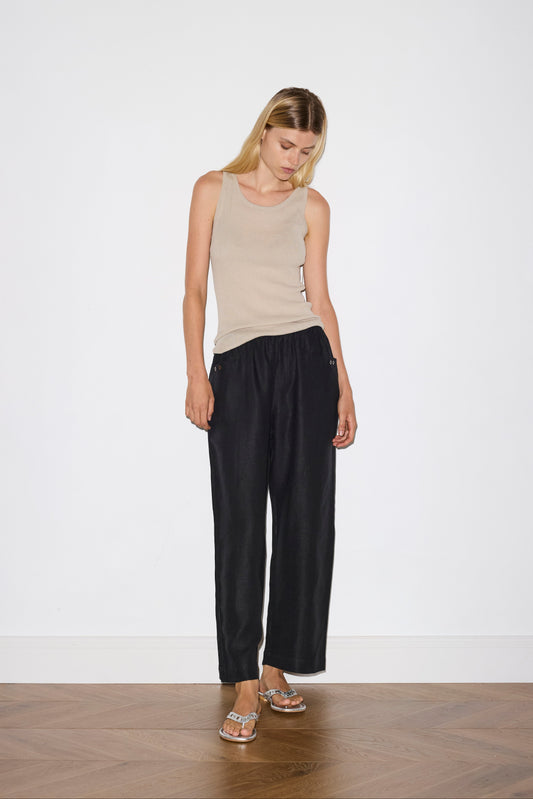 blonde haired female model wears black an ankle length, straight leg pant features a soft minimal elastic waist with front seam pockets and button detail, paired with a beige a minimalist tank-style top featuring raw edge detailing and an asymmetrical tie strap design for a unique and adjustable fit. made with a lightweight ribbed texture