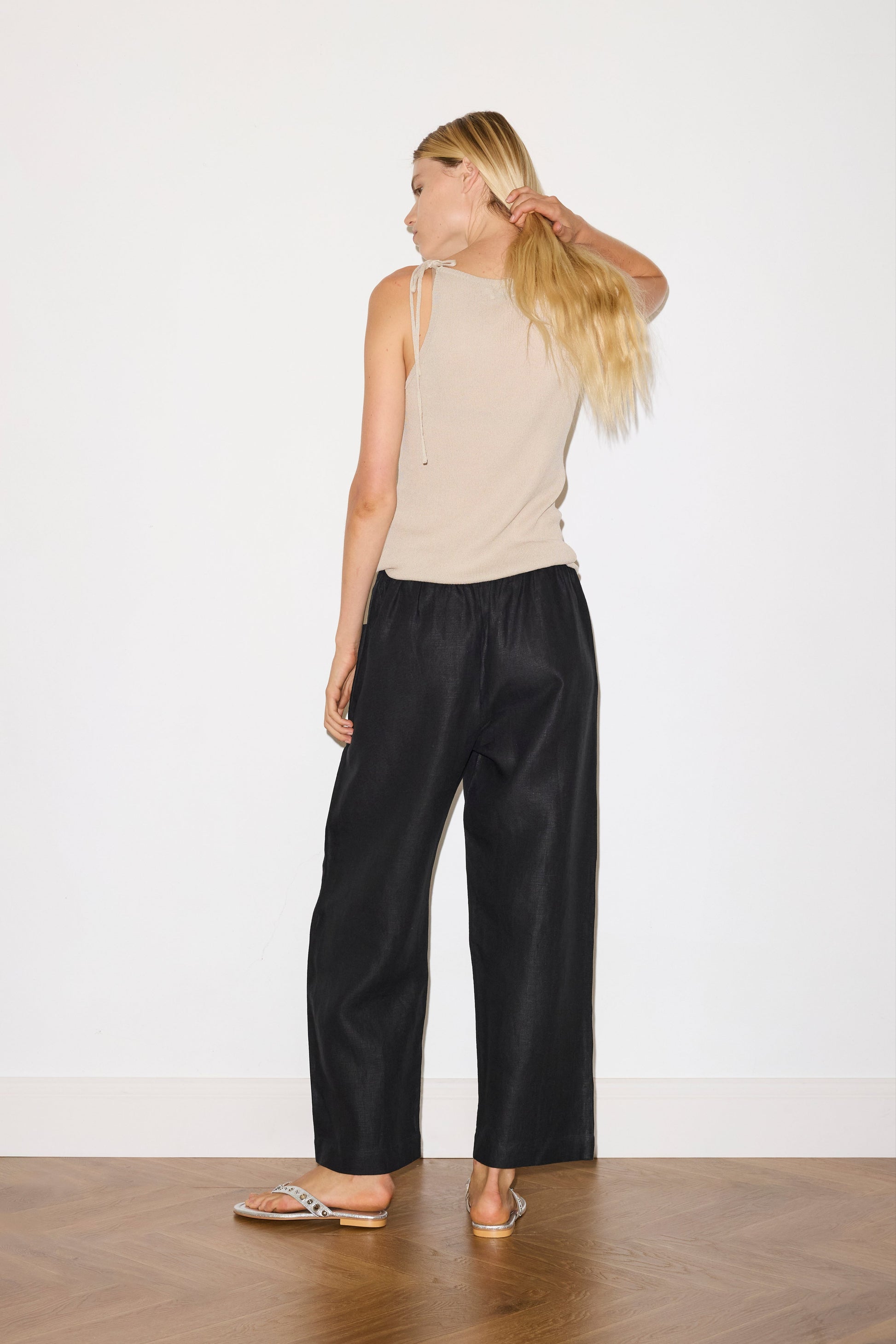 blonde female model wears a beige minimalist tank-style top featuring raw edge detailing and an asymmetrical tie strap design for a unique and adjustable fit. made with a lightweight ribbed texture, worn with a black ankle length, straight leg pant features a soft minimal elastic waist with front seam pockets and button detail